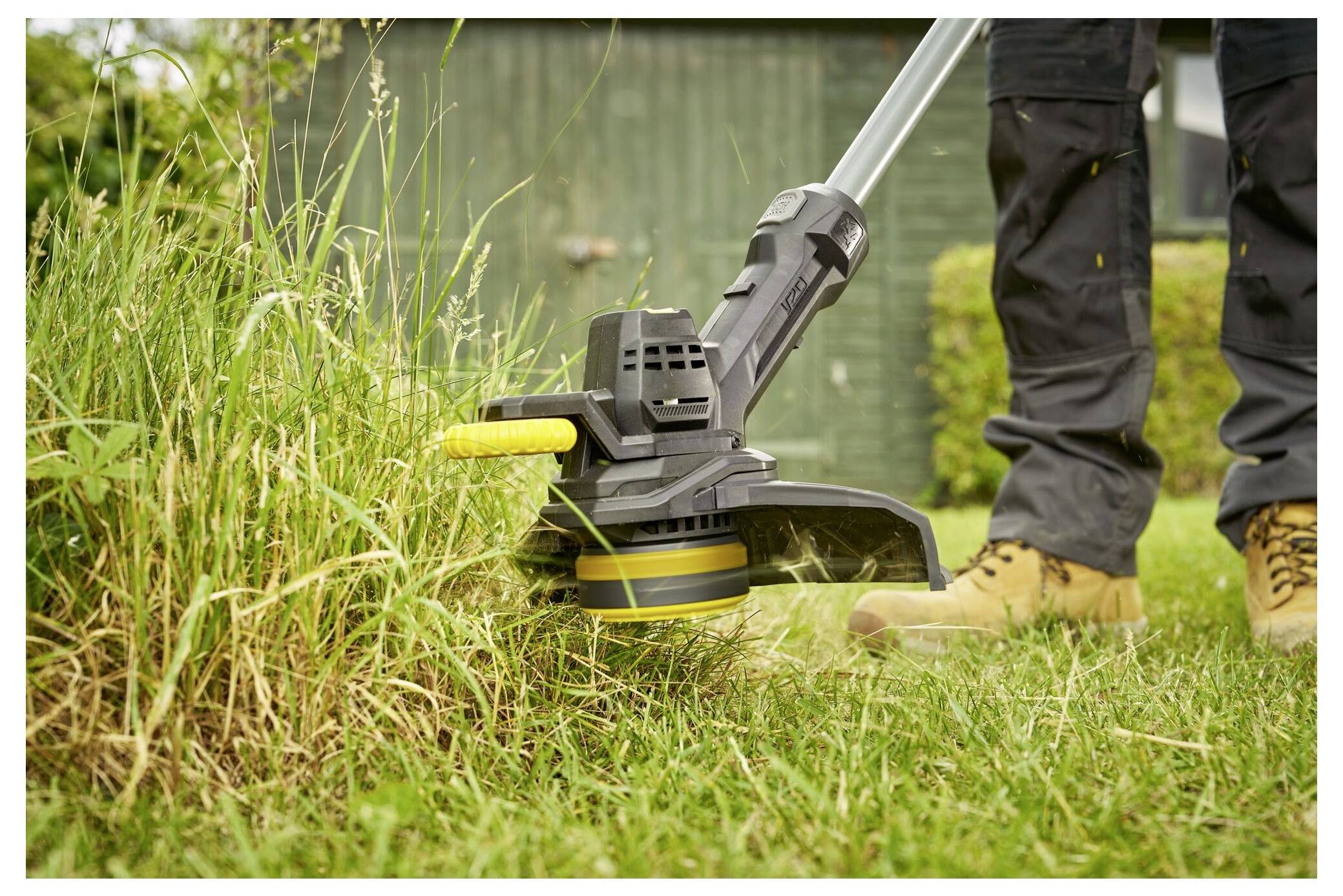 A person trims overgrown grass with a yellow and black electric lawn trimmer in a garden, wearing brown boots and dark pants.