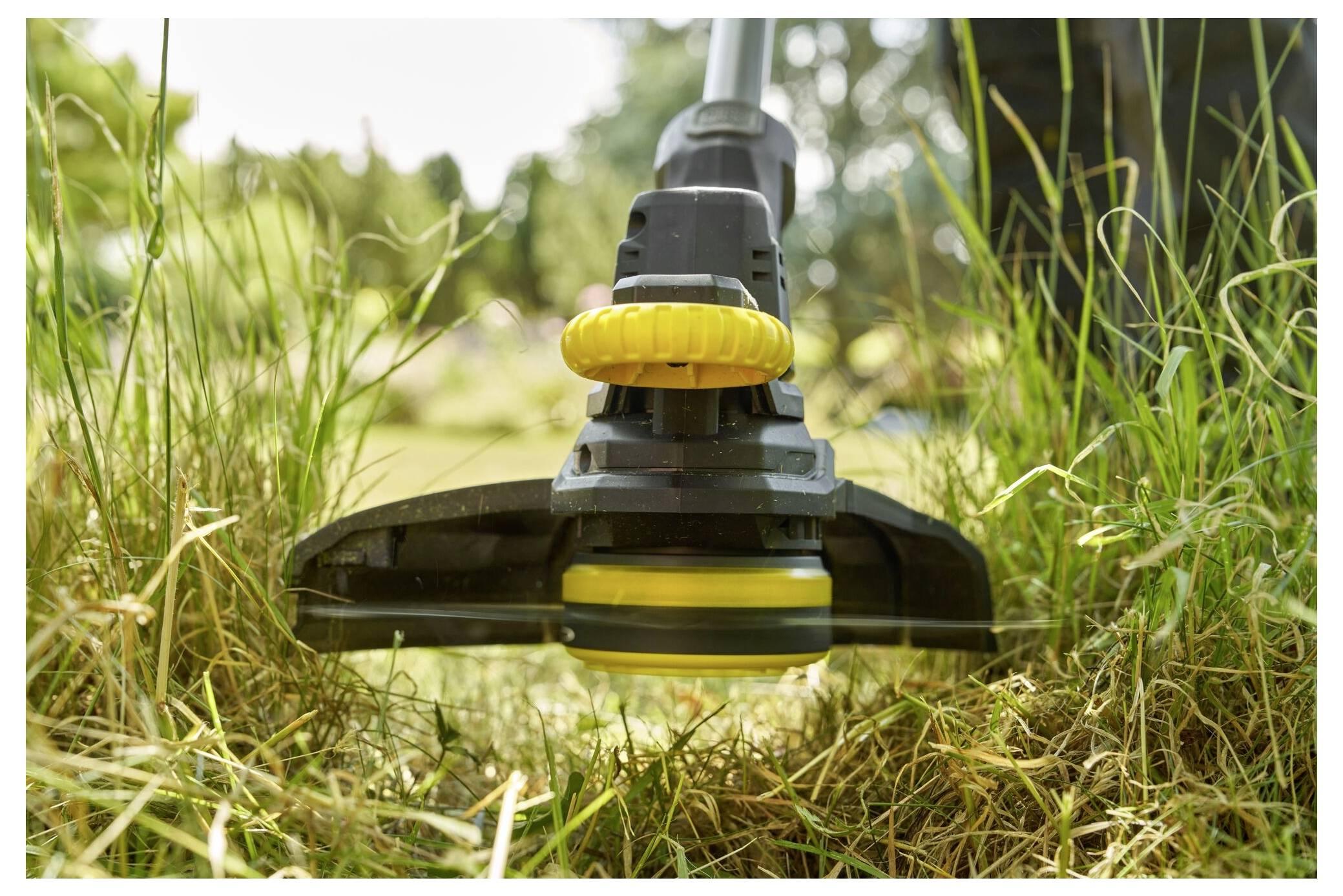Close-up of a grass trimmer cutting grass, focusing on the rotating blades. The background shows a blurred garden setting.