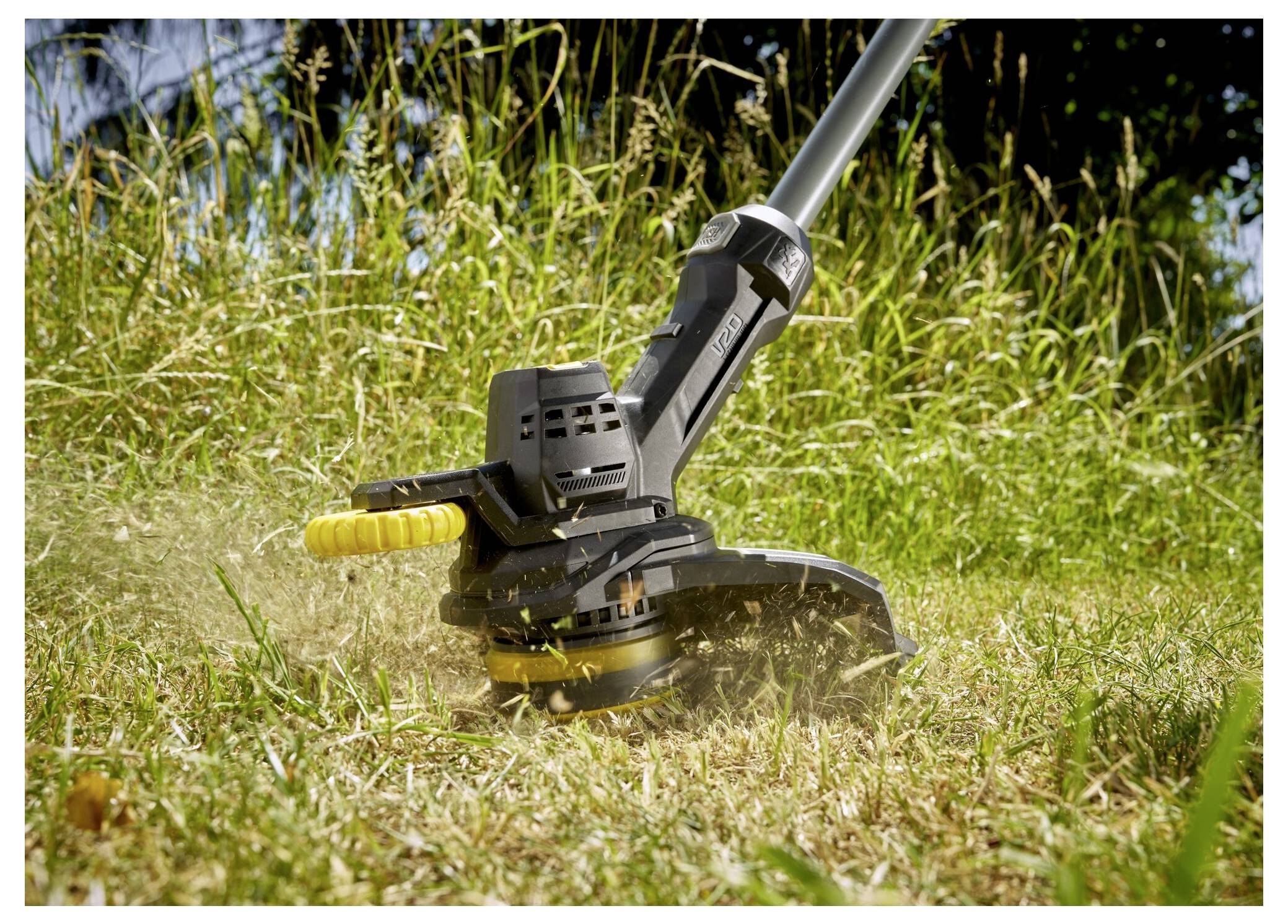 A black and yellow grass trimmer cutting through overgrown grass in a garden, with trees in the background.
