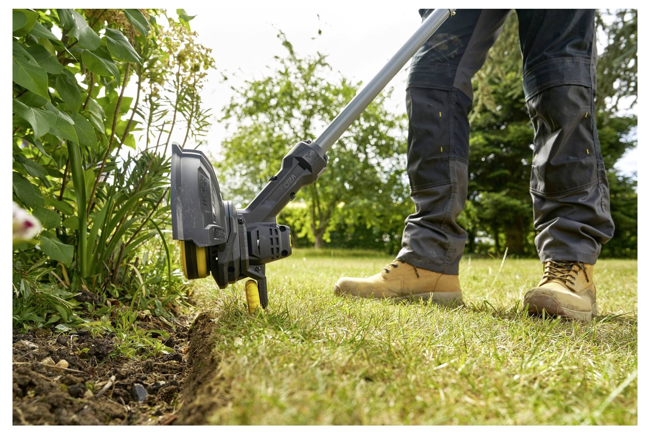 A person wearing work boots uses a garden edger to trim the edge of a grass lawn next to a garden bed with green plants.