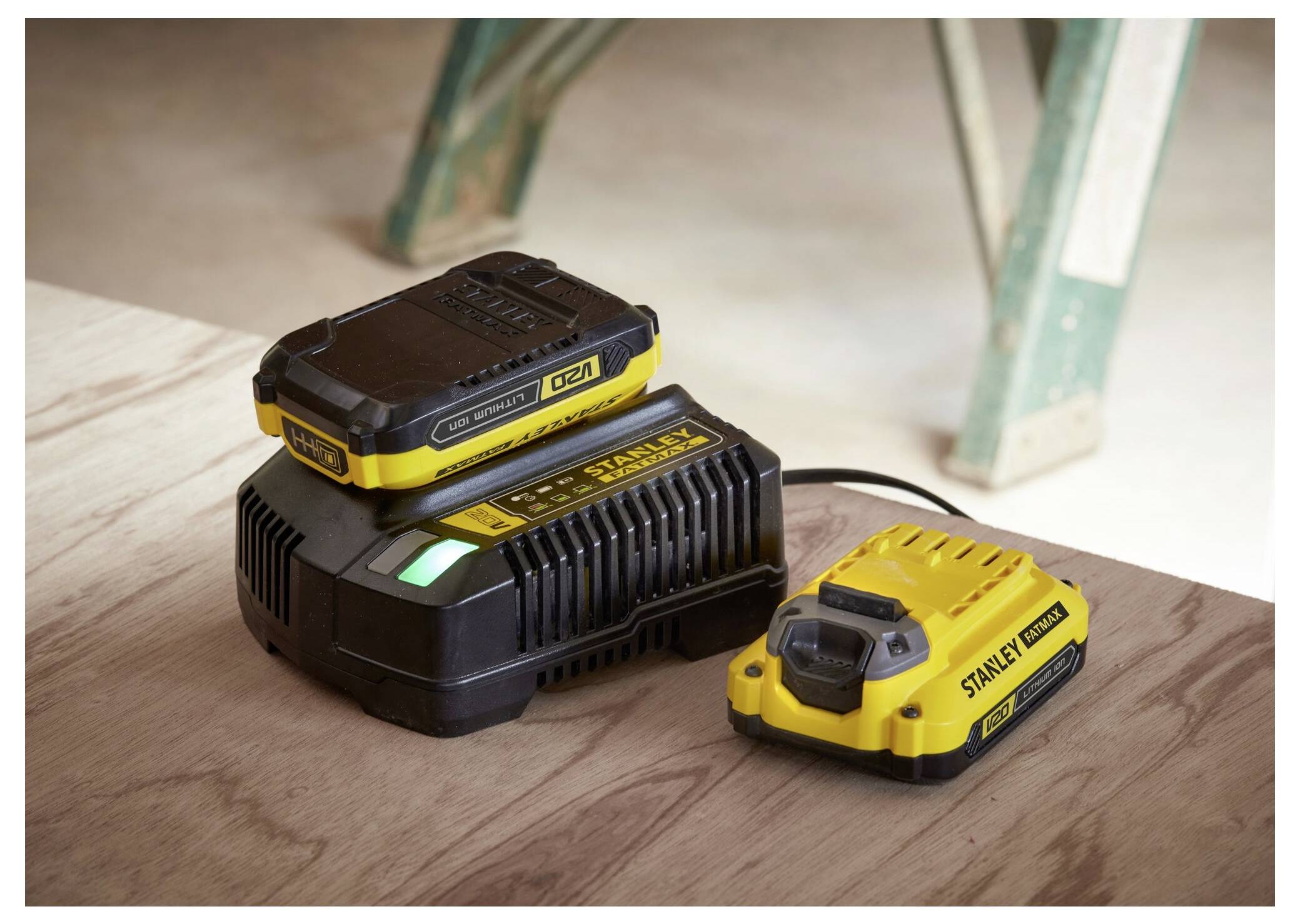 'A yellow and black Stanley battery charger with a charged battery is placed on a wooden surface, next to a pair of ladder legs.'