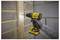 A person using a yellow and black cordless drill to make a hole in a wooden plank against a brick wall.