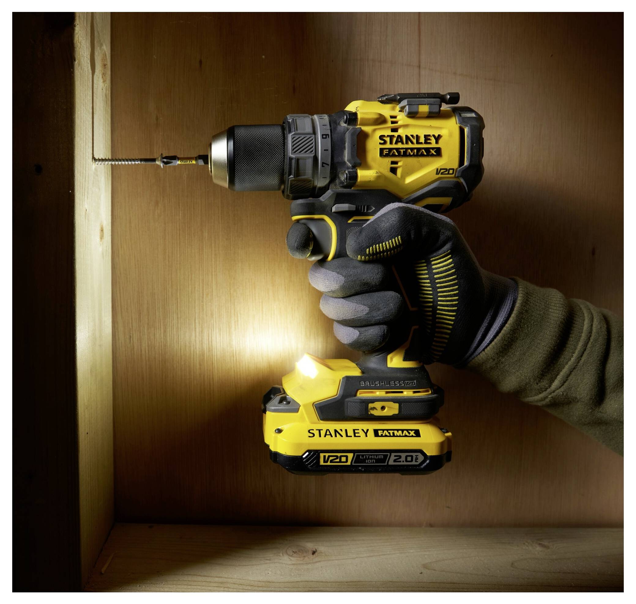 A hand wearing a glove uses a yellow Stanley Fatmax V20 cordless drill to drive a screw into wooden paneling.