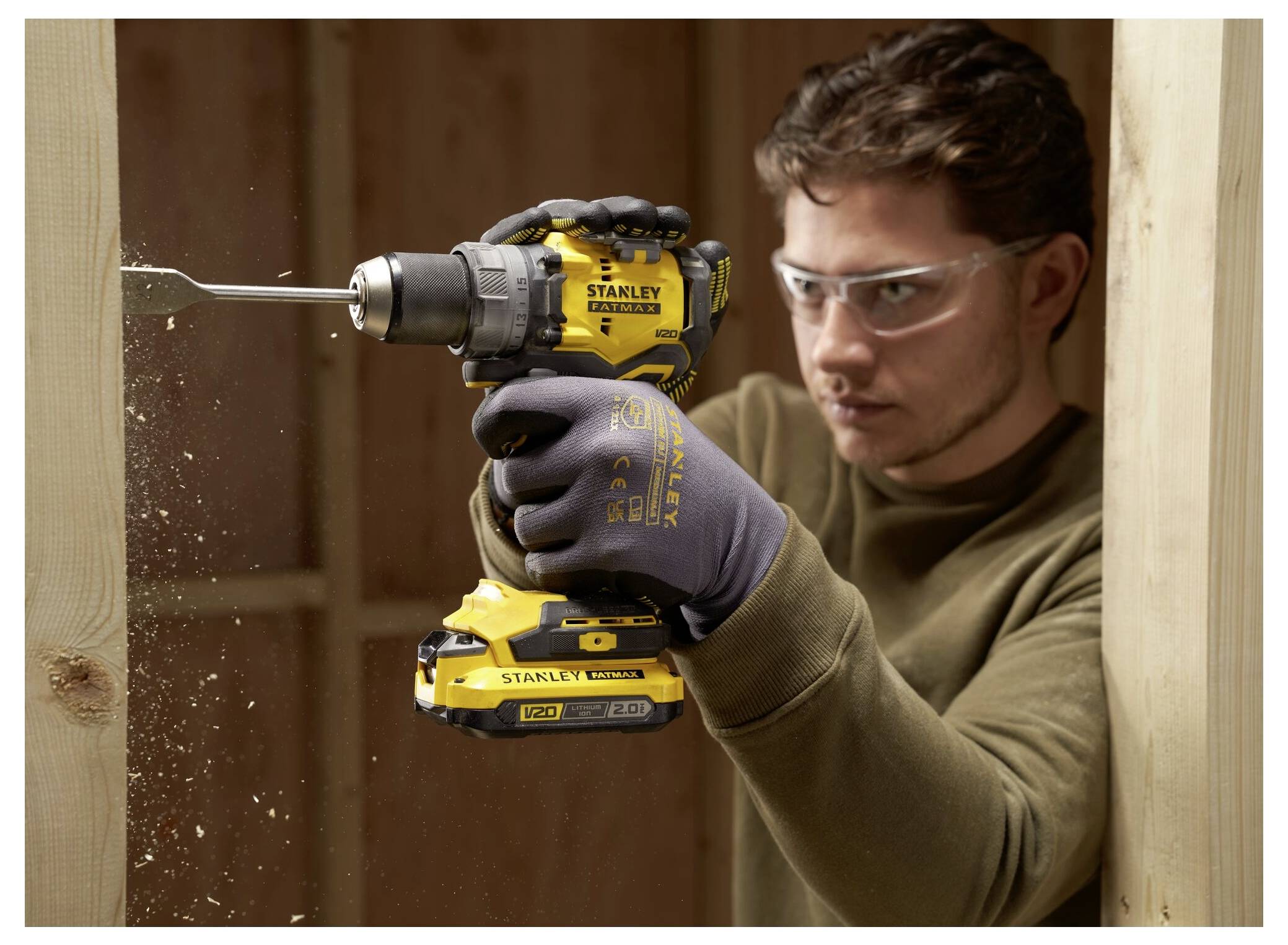 A person wearing safety goggles and gloves uses a yellow Stanley electric drill to bore a hole into a wooden plank, creating wood shavings.
