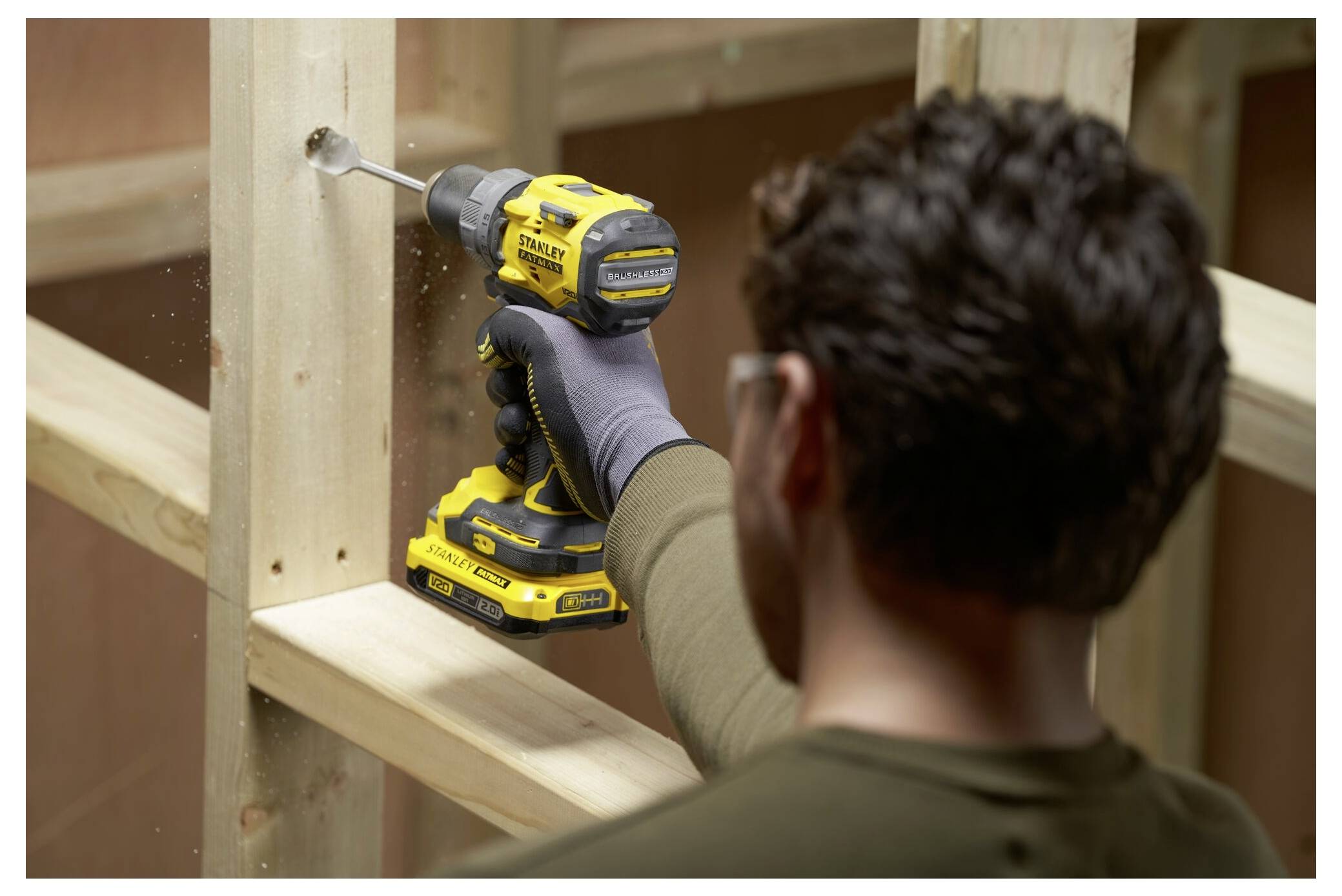 A person is using a yellow cordless drill to make a hole in a wooden beam within a construction frame, wearing safety glasses and gloves.