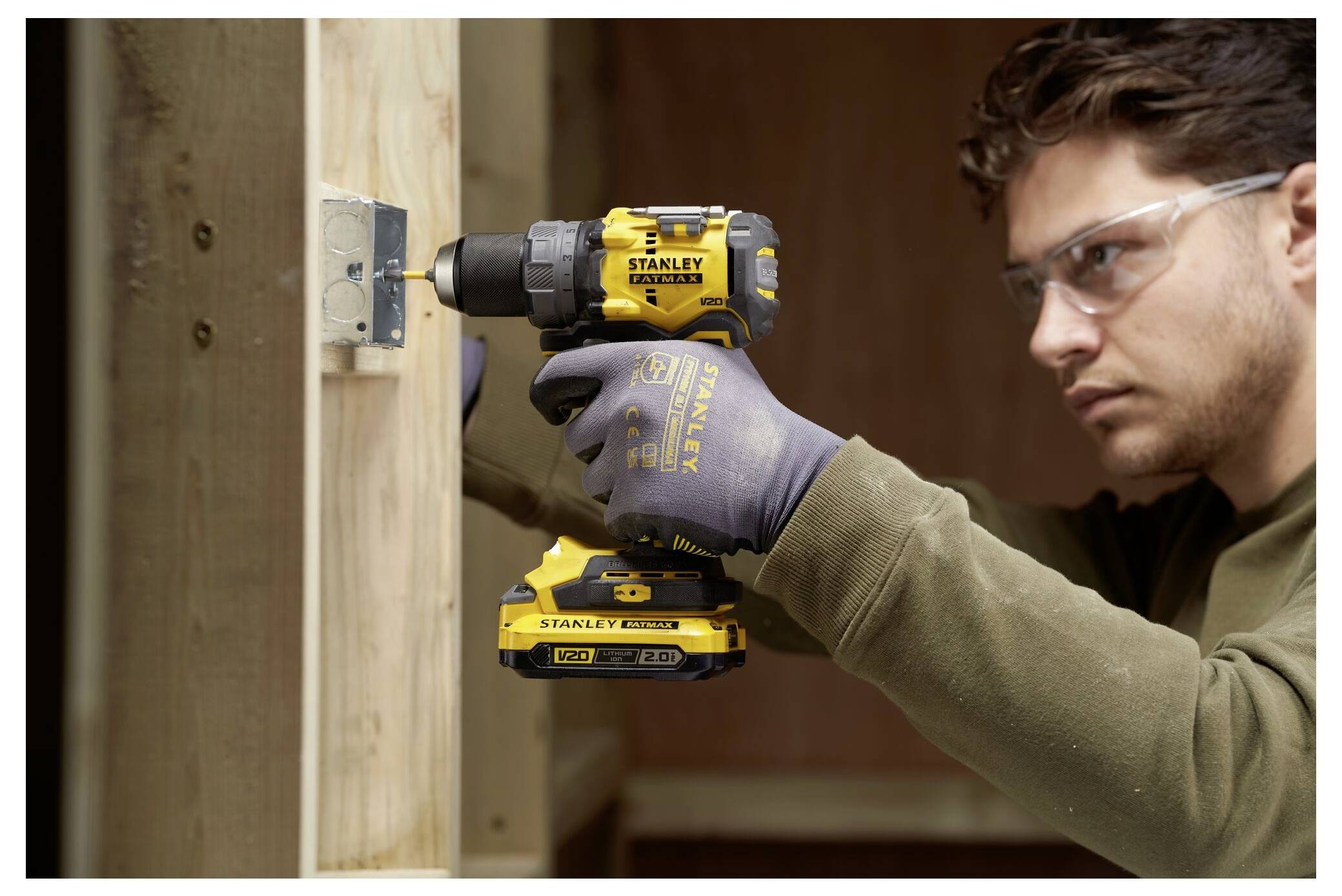 A person uses a yellow and black power drill to secure a metal bracket to a wooden structure, wearing safety glasses and gloves.