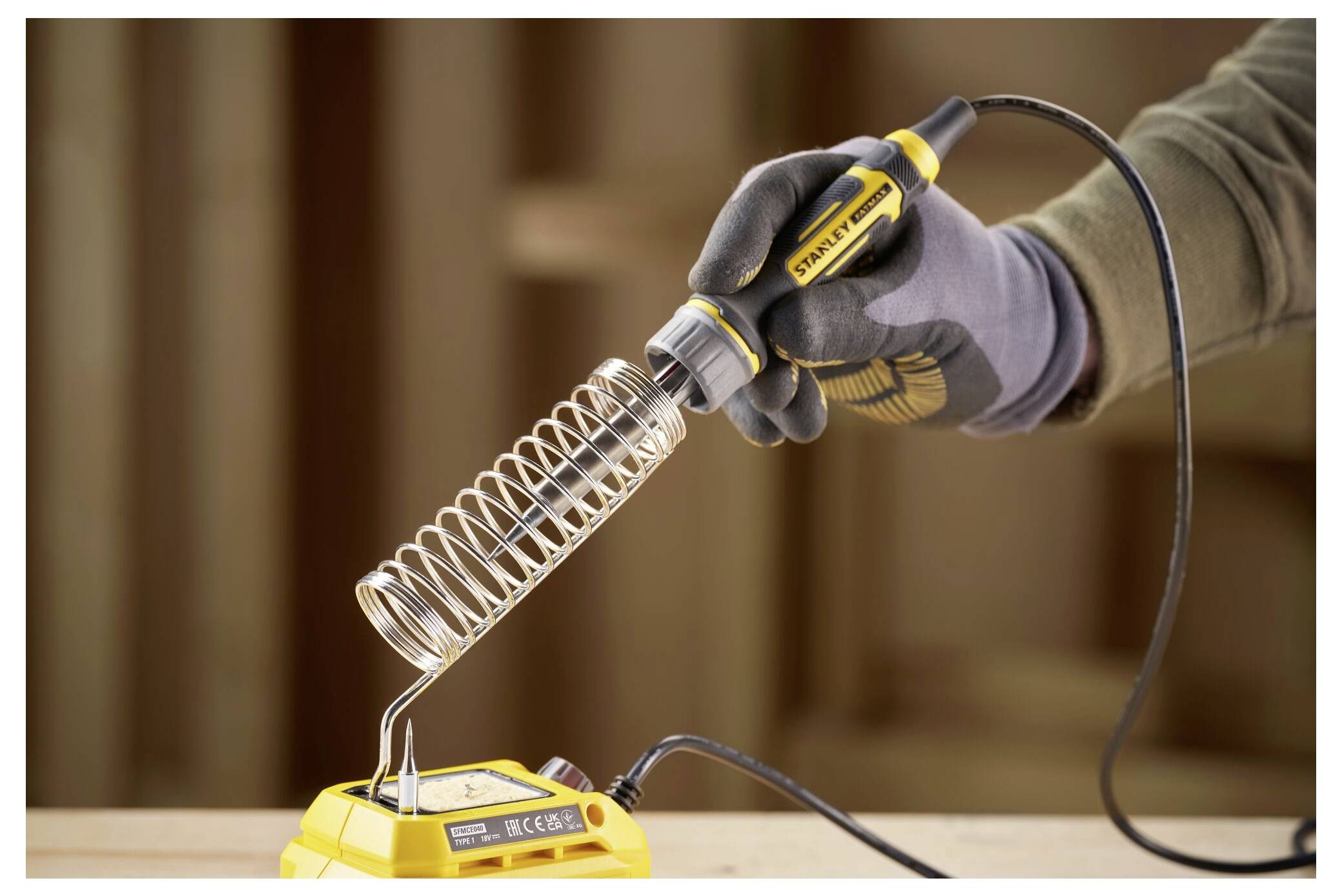 A gloved hand holds a soldering iron above its stand on a workbench, with a wooden background.