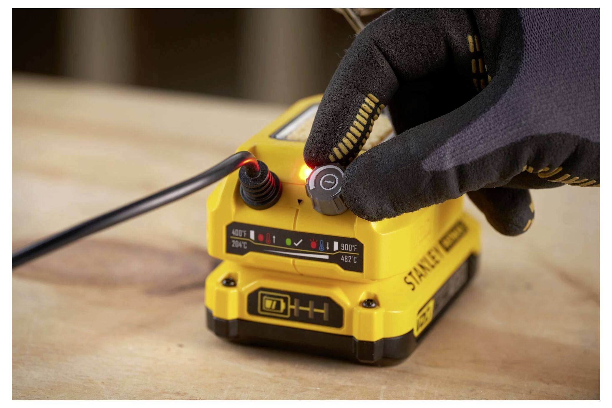 A gloved hand adjusts a dial on a yellow power tool battery with a digital display showing charging status on a wooden surface.