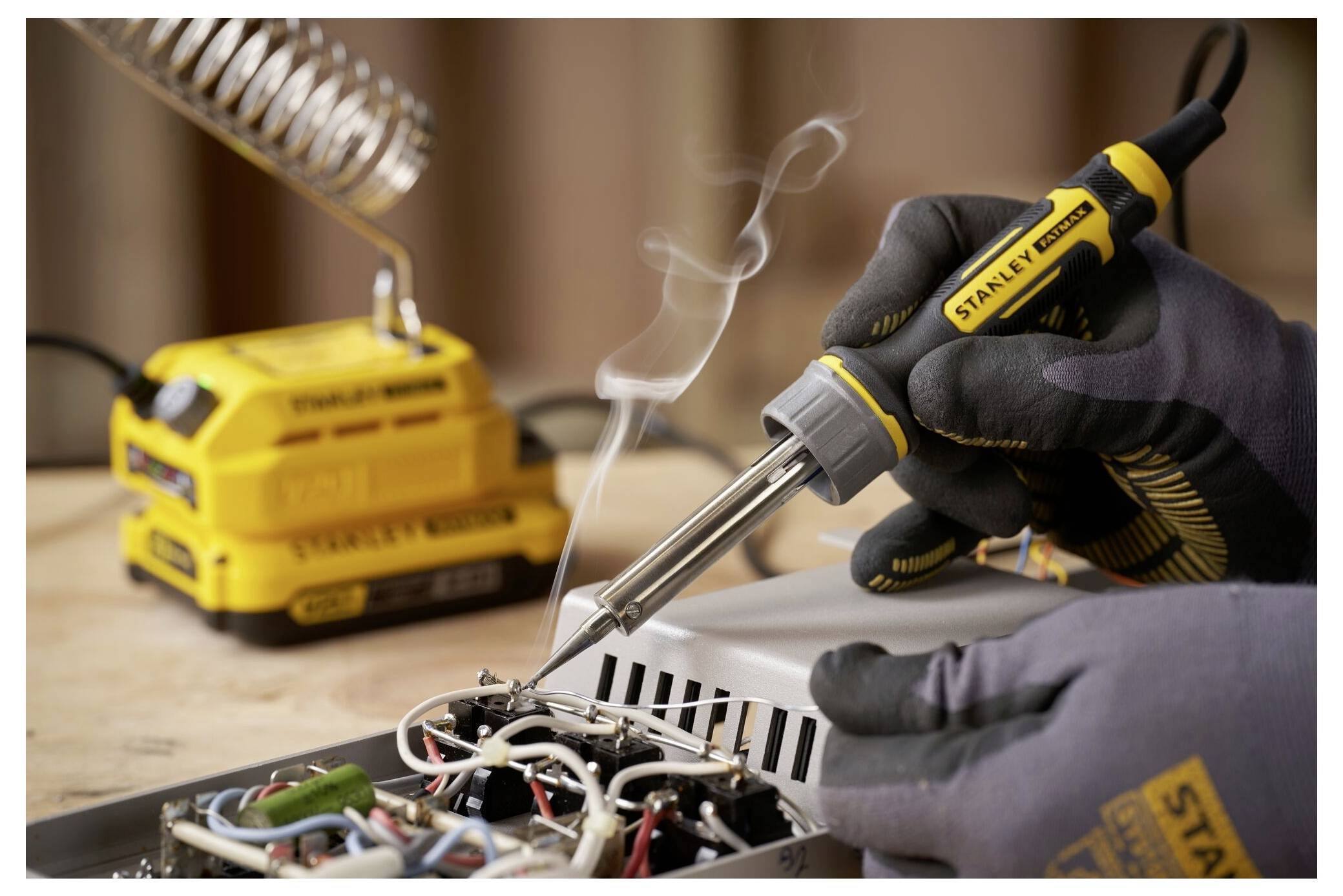 A person wearing gloves is using a soldering iron to work on electronic components, with a soldering station and smoke visible.