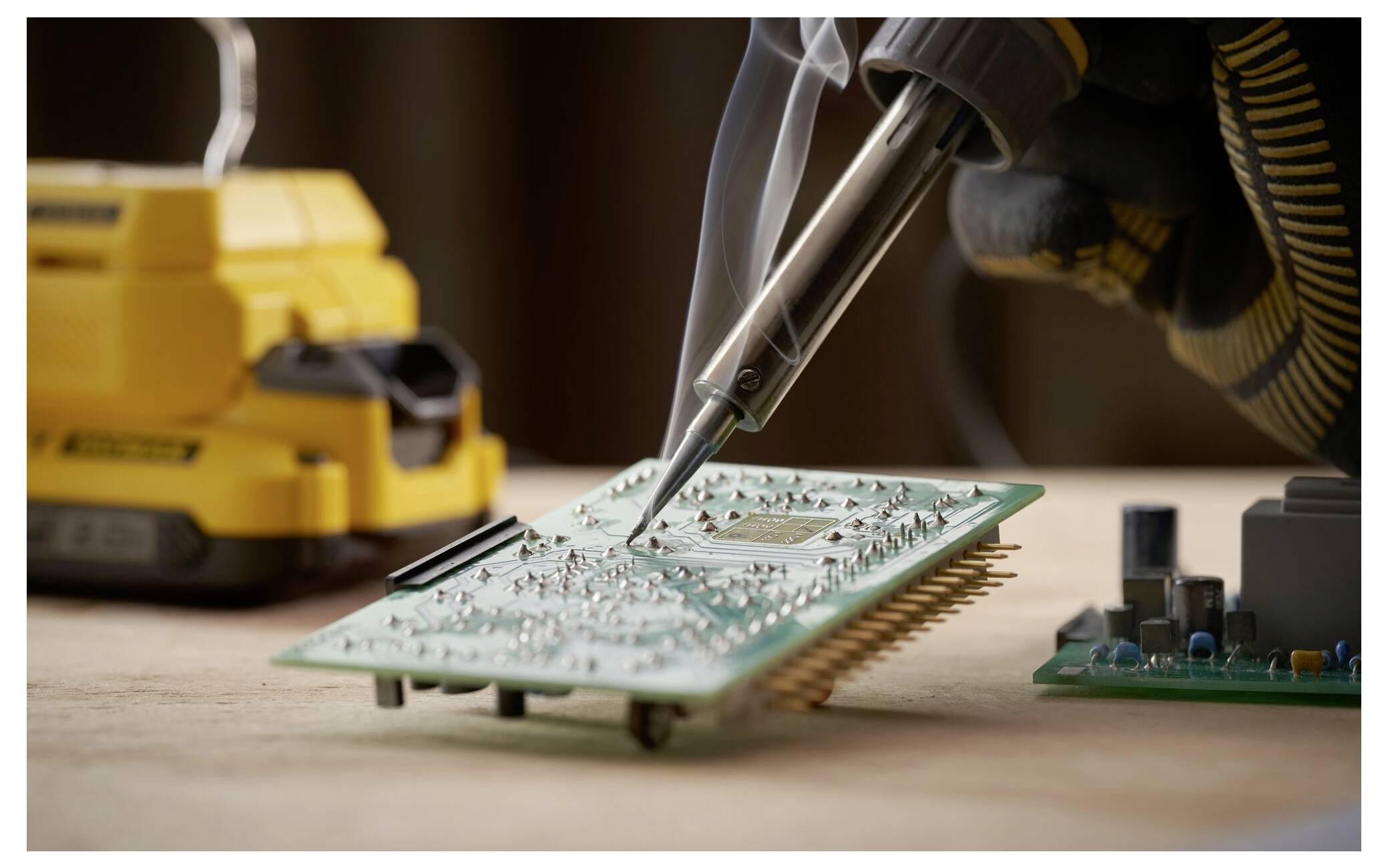A gloved hand solders a component on a green circuit board. A yellow soldering station and another circuit board are in the background.