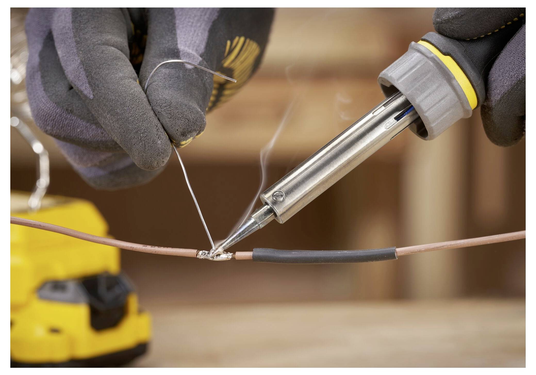 Close-up of gloved hands using a soldering iron to connect wires, with smoke rising. A yellow tool is blurred in the background.