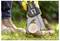 A person in work boots uses a hand-held electric chainsaw to cut a log on grassy ground. The tool is branded and yellow.