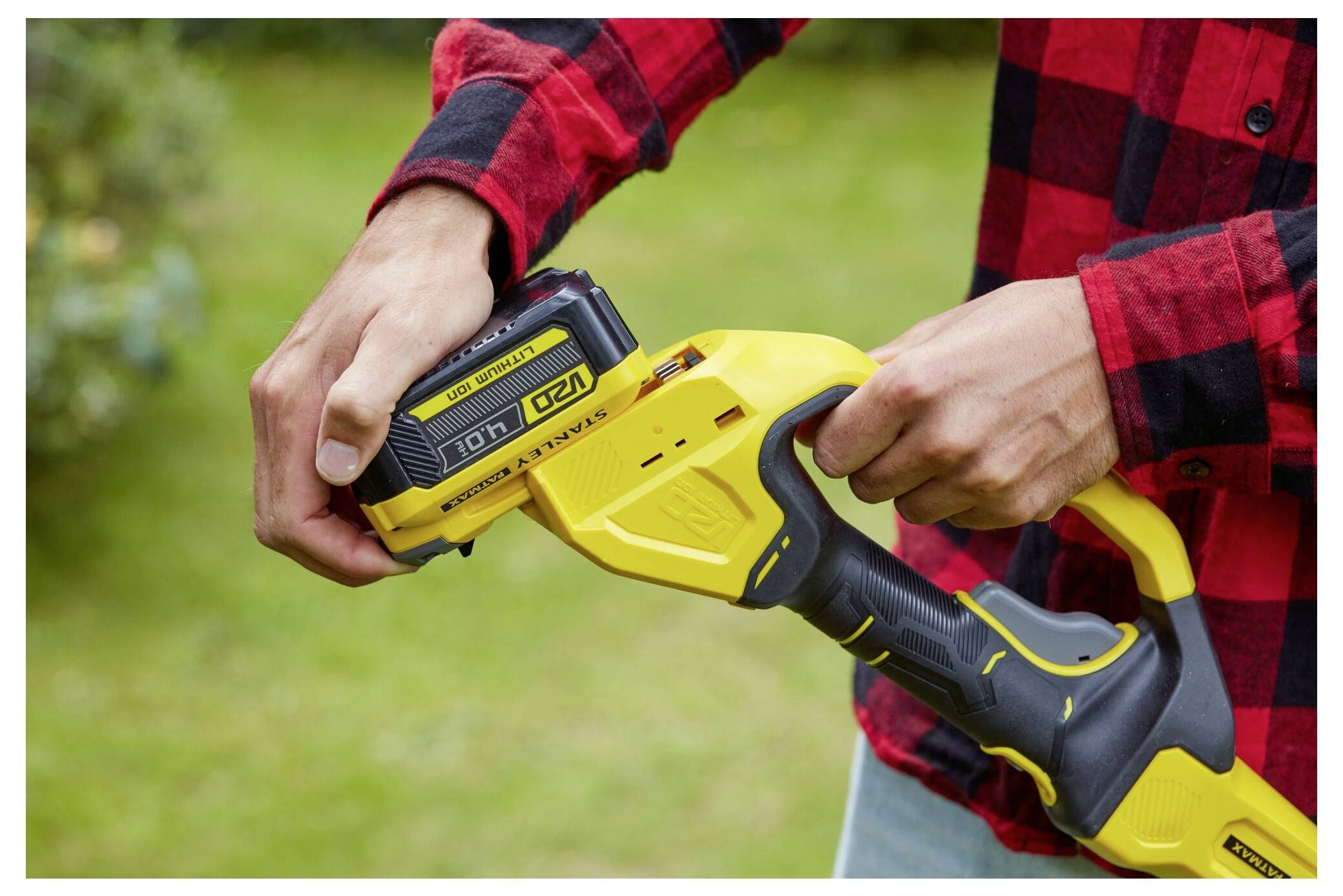 A person in a red plaid shirt inserts a 10Ah 20V lithium battery into a yellow cordless power tool, outdoors on a grassy lawn.