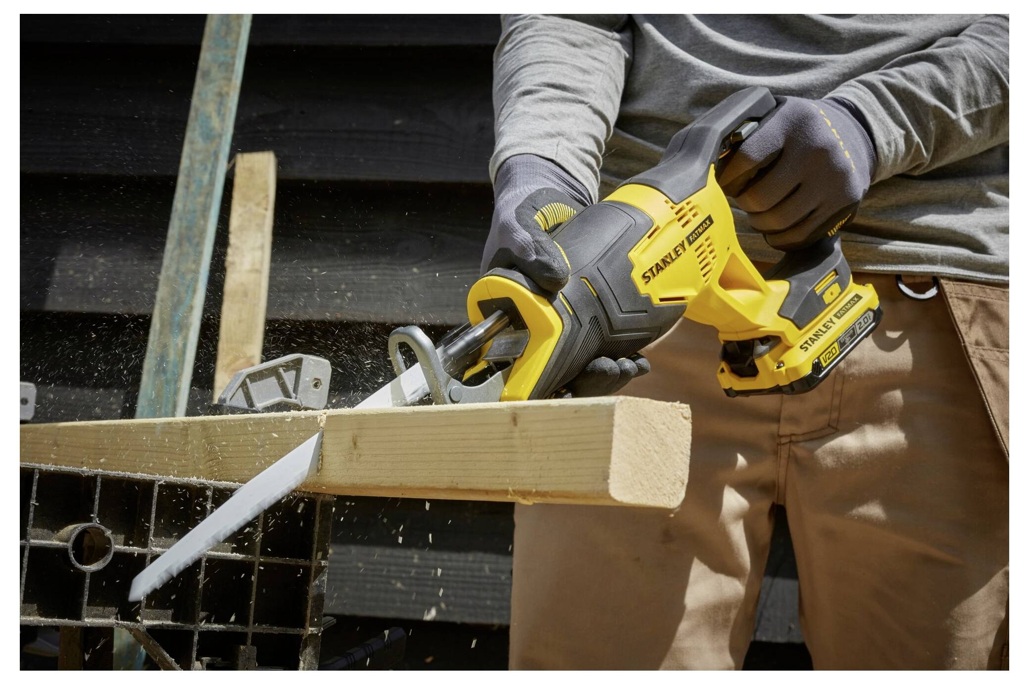 A person cutting a wooden plank with a yellow reciprocating saw on a workbench, wearing gloves and brown pants.