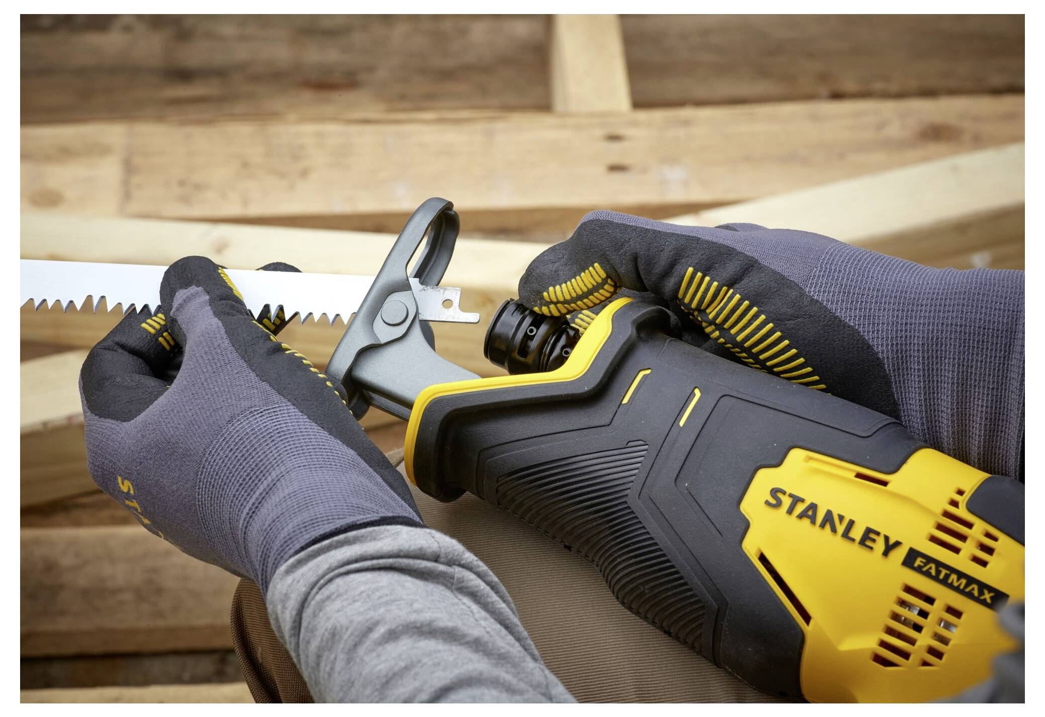 'Hands wearing gloves hold a Stanley Fatmax power saw, adjusting its blade. Wooden planks are visible in the background.'