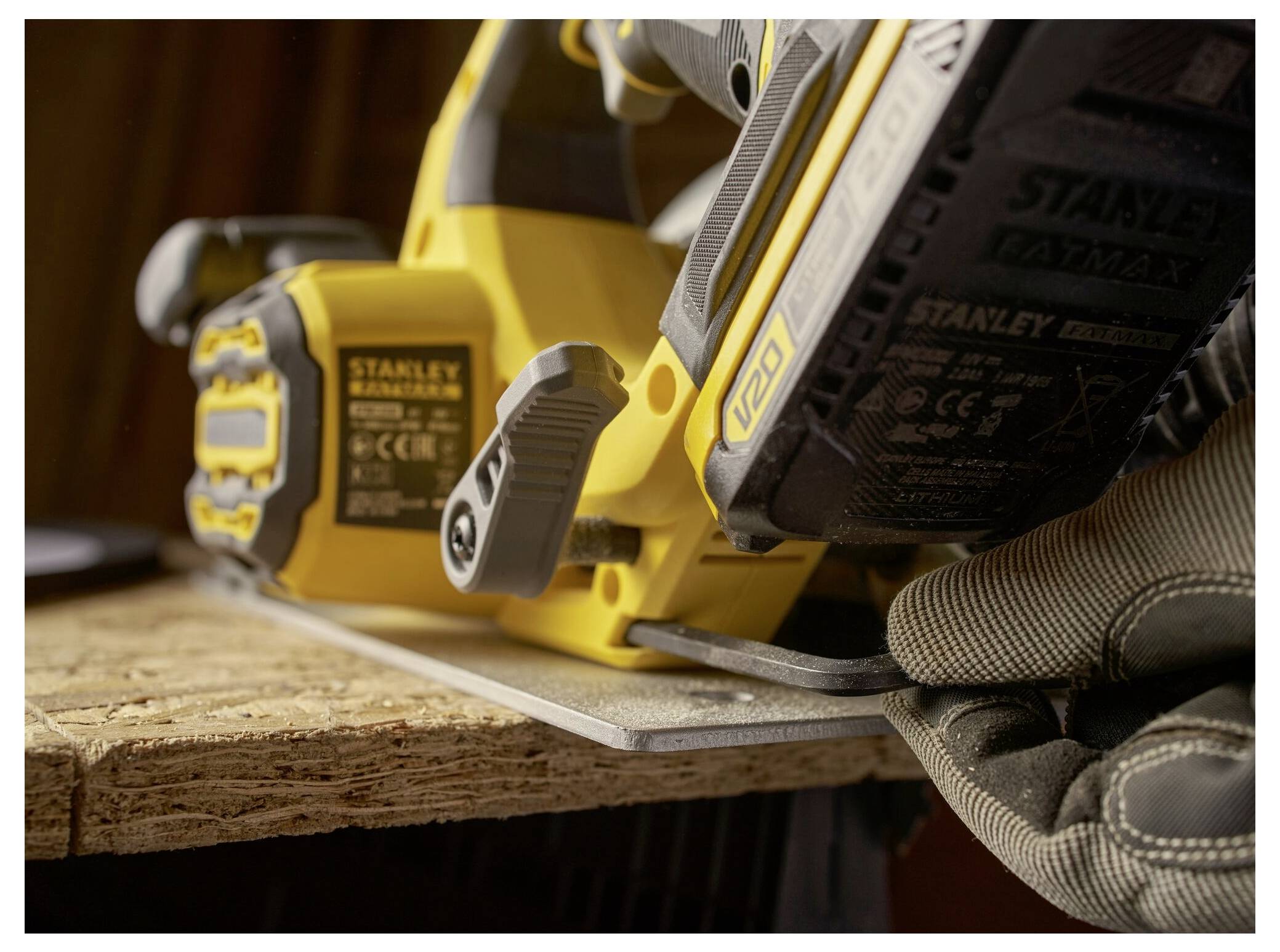 A close-up of a person using a yellow Stanley power tool to cut through a wooden plank, wearing protective gloves for safety.