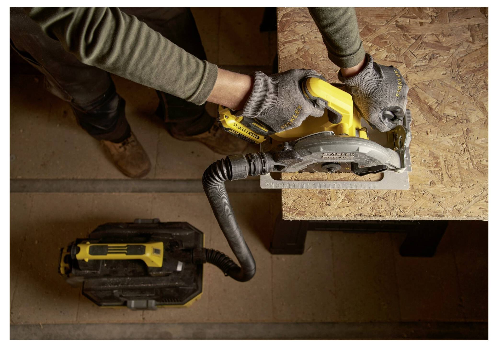 A person using a circular saw to cut a wooden board, with a vacuum hose attached to collect dust. Safety gloves and a stable setup are in use.