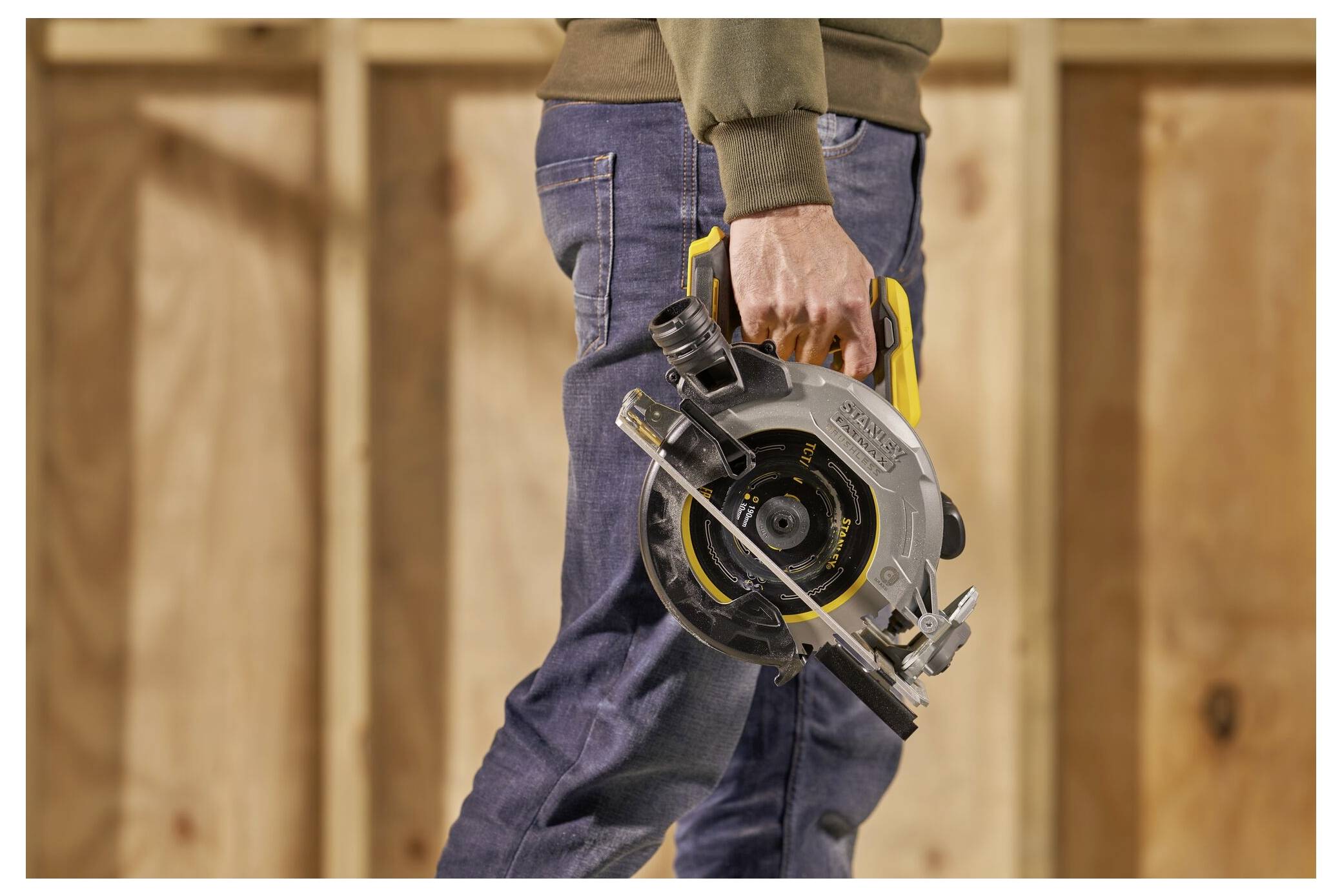 A person holding a circular saw by its handle, wearing jeans and a sweater, standing against a wooden wall backdrop.