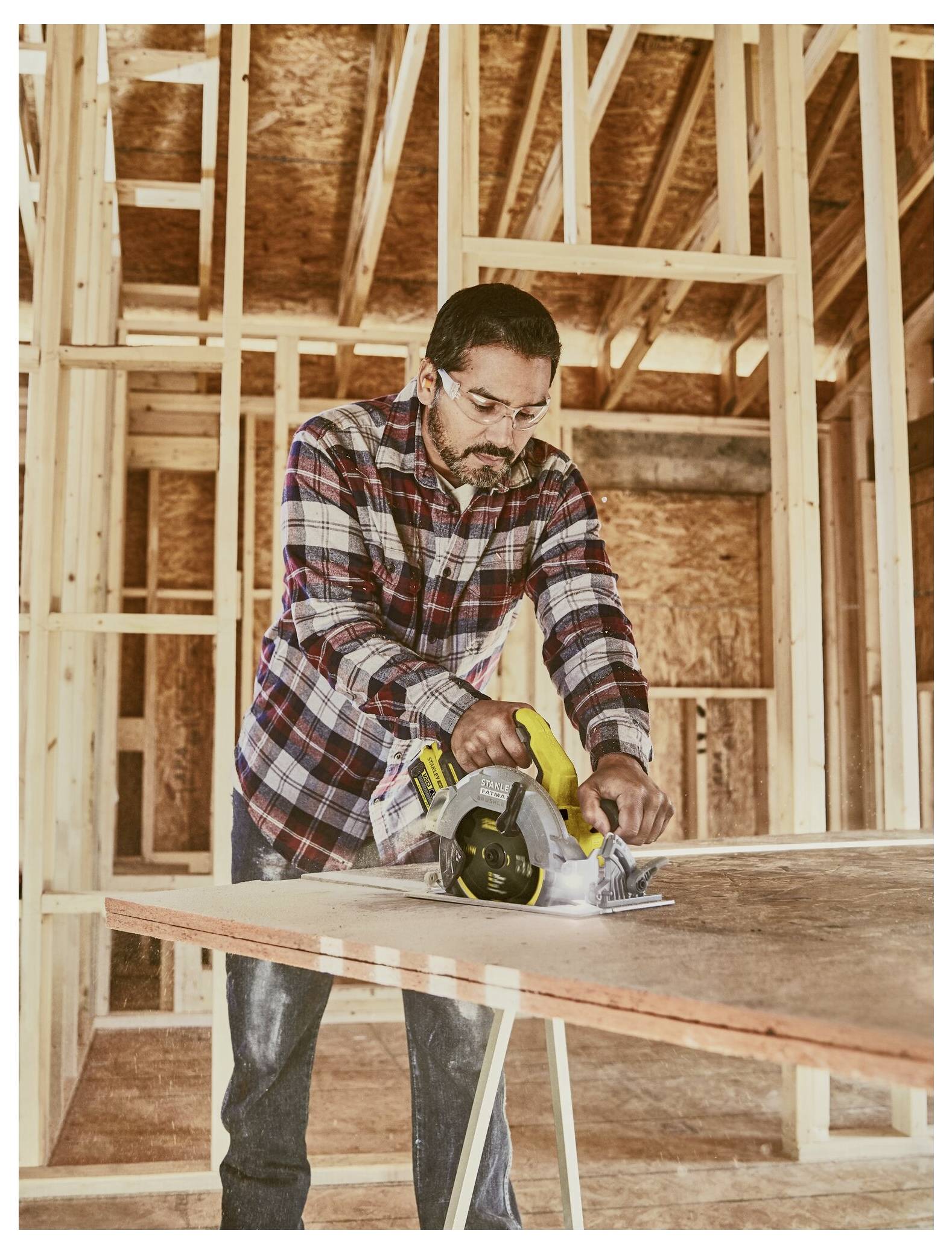 A person wearing safety glasses uses a circular saw to cut a wooden board in a partially constructed wooden building interior.