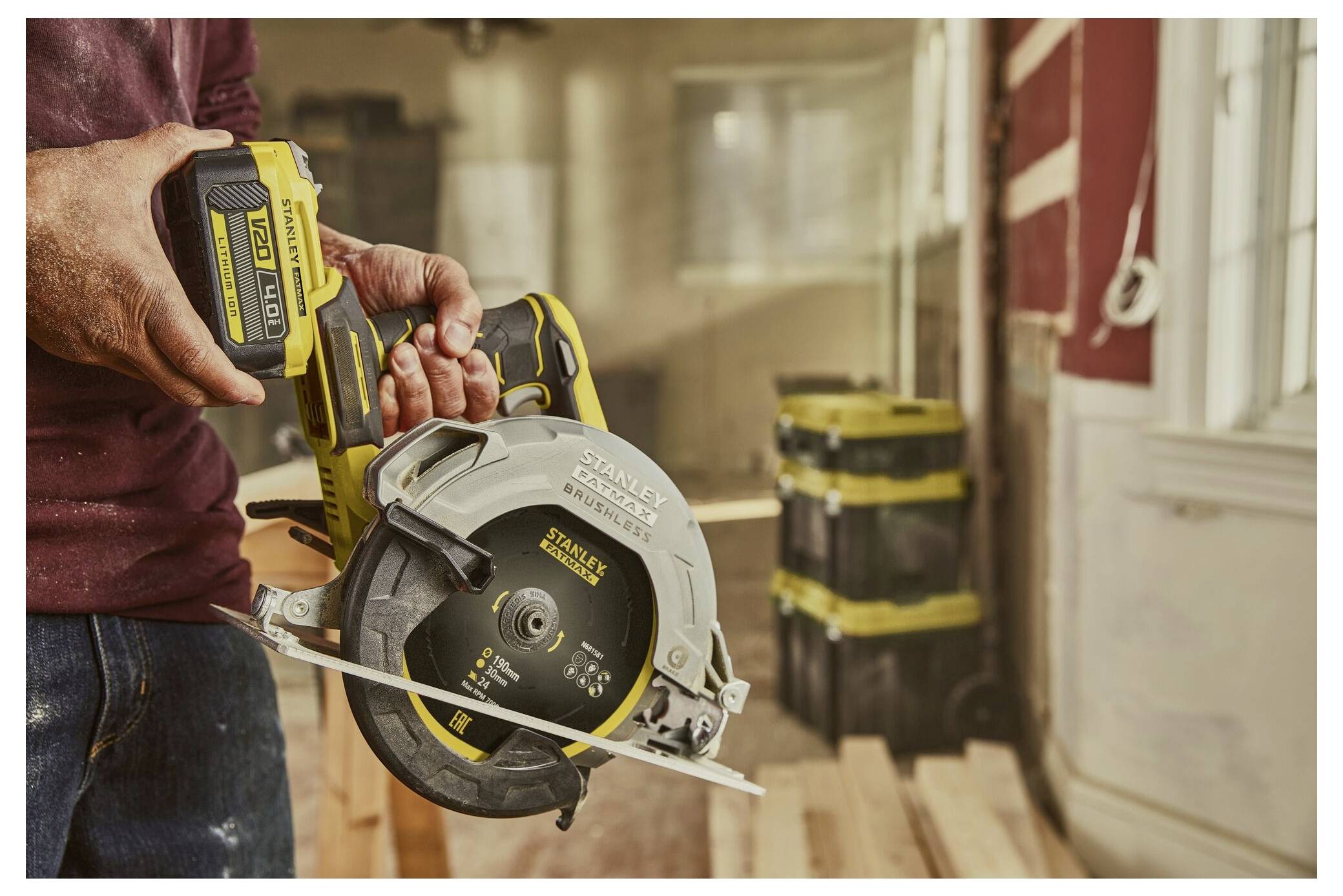 A person holds a cordless circular saw in a workshop with stacked toolboxes and wooden planks in the background, suggesting construction work.