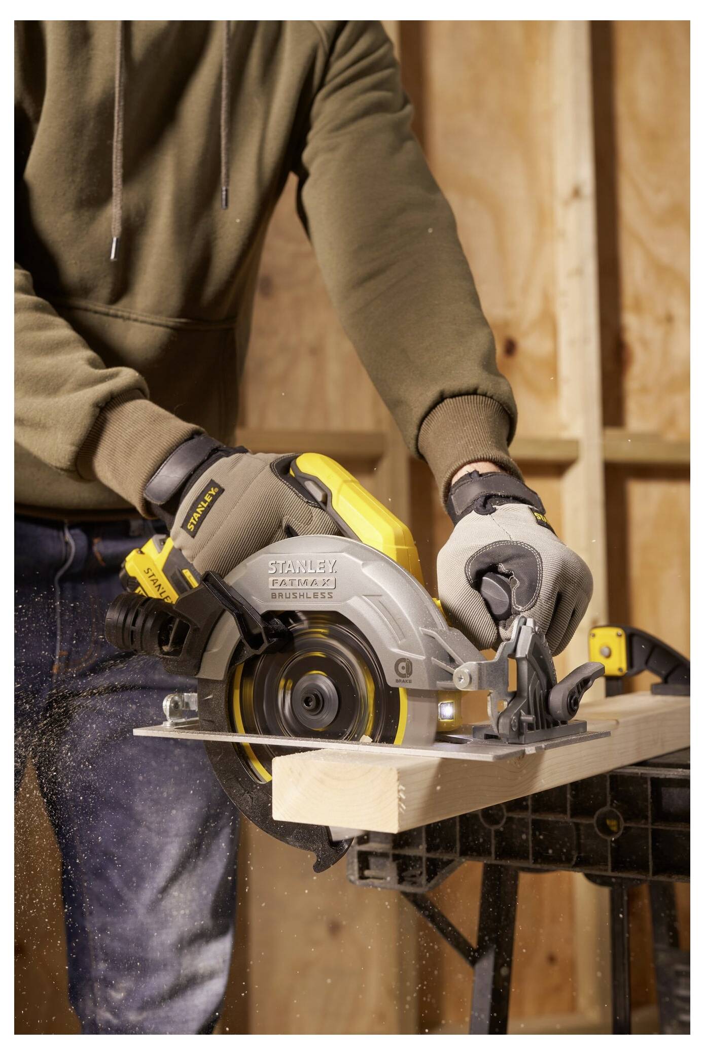 A person using a circular saw to cut a wooden plank in a workshop. The person is wearing safety gloves and a hoodie.