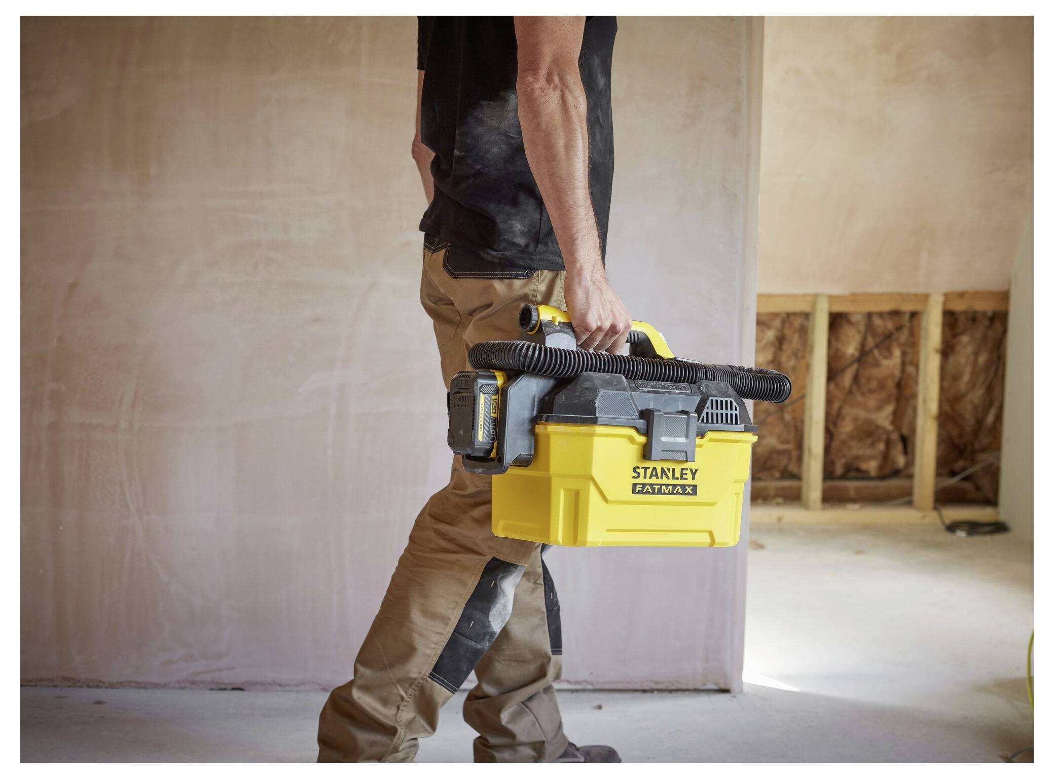 A person carries a yellow 'Stanley FatMax' toolbox in a construction area. Walls are partially finished, and tools are attached to the box.