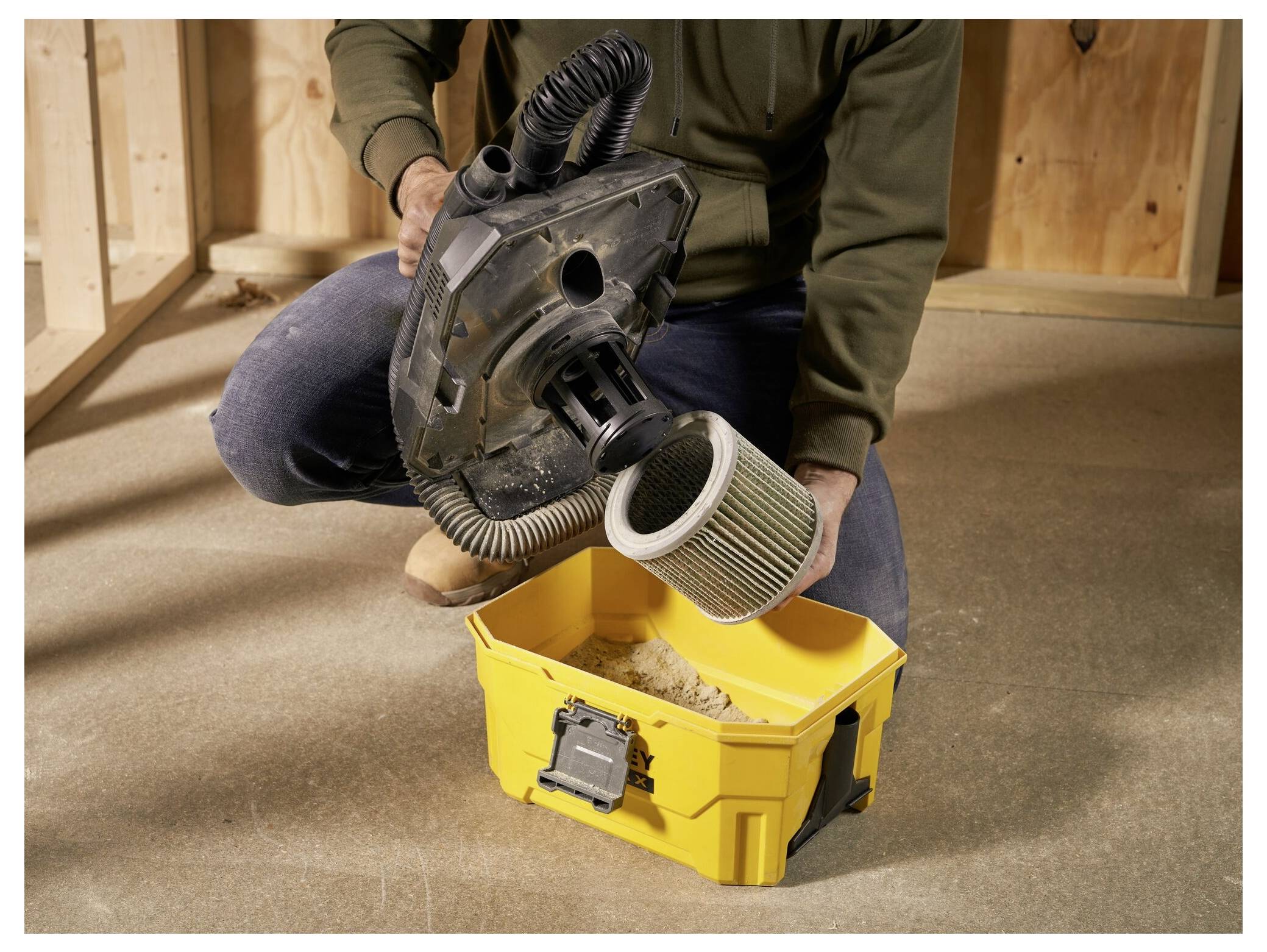 A person in work clothes kneeling on a concrete floor empties a cylindrical vacuum filter into a yellow container.