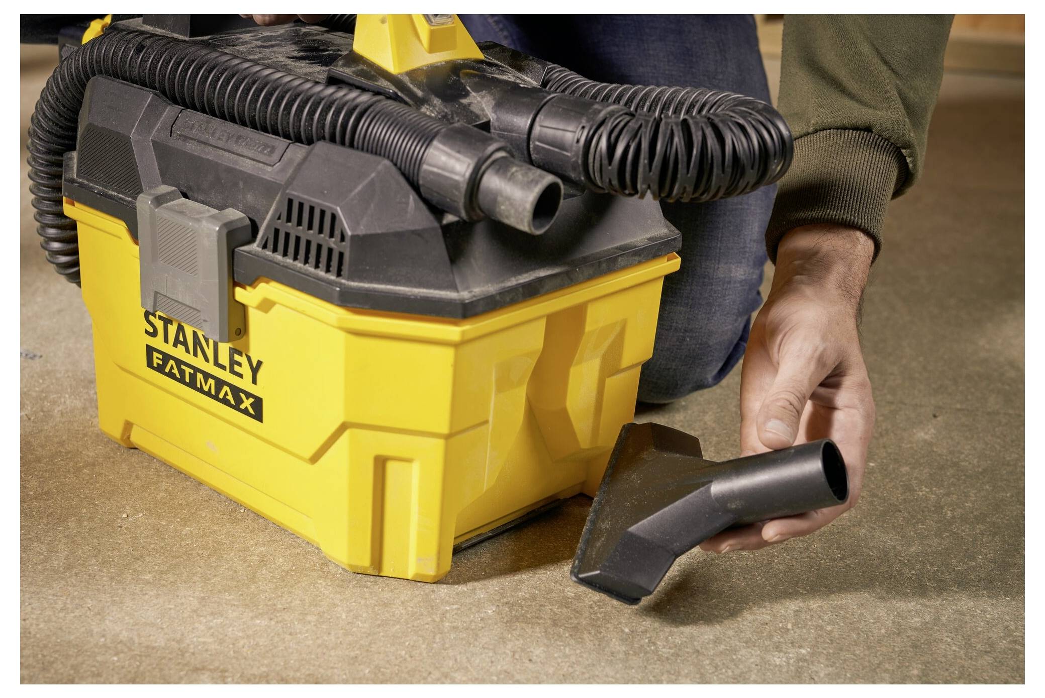 A person uses a yellow 'Stanley Fatmax' vacuum cleaner on a concrete floor, holding a black hose attachment close to the ground.