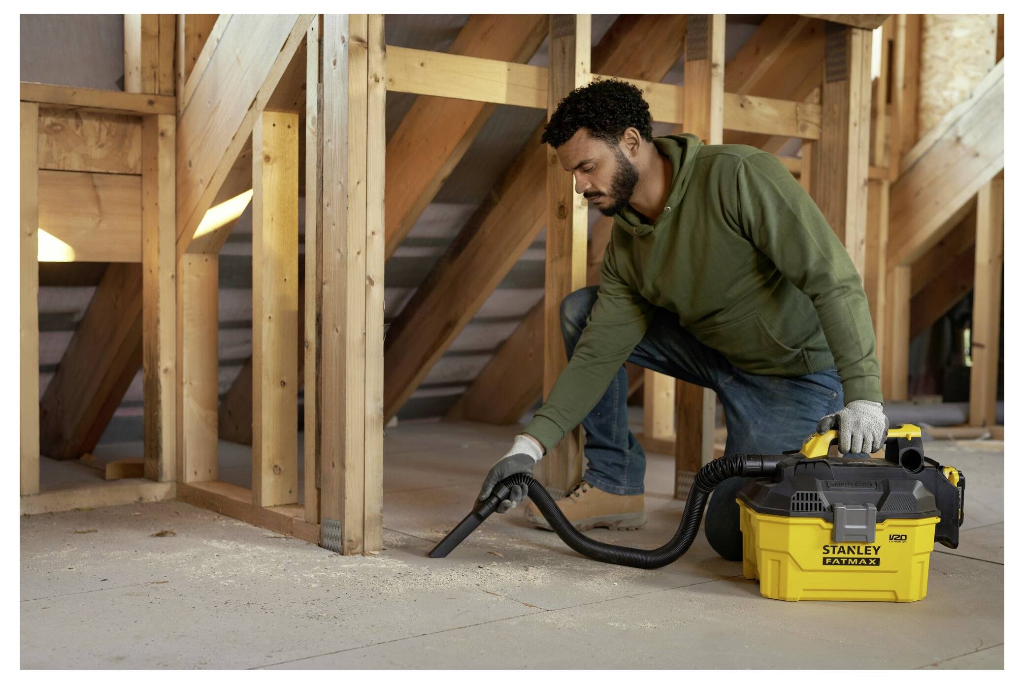 A man cleans construction debris in an unfinished attic using a Stanley wet/dry vacuum. He wears casual work clothes and gloves.