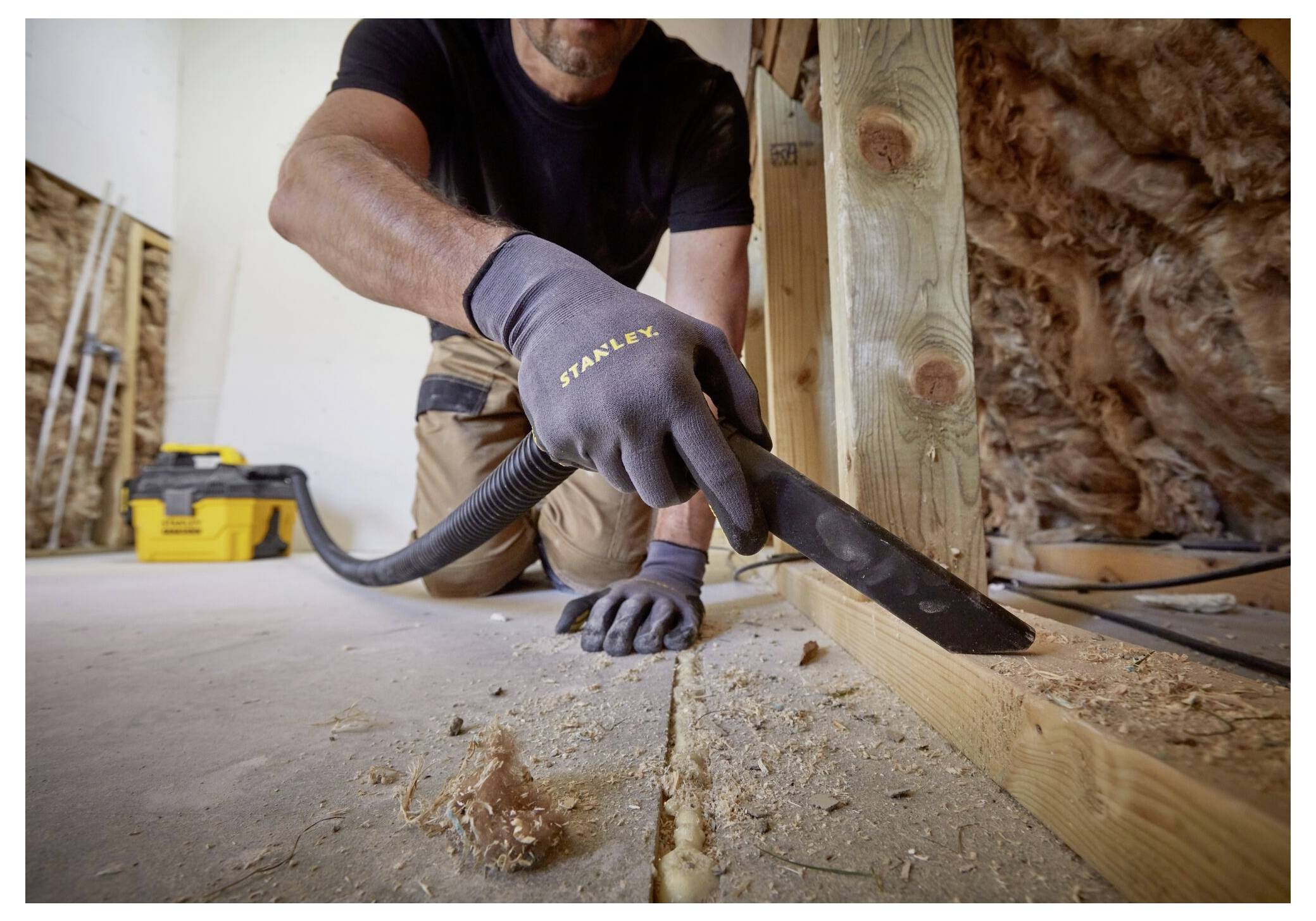 A person cleans sawdust from a construction site floor using a vacuum. They wear protective gloves and kneel beside wooden framing.