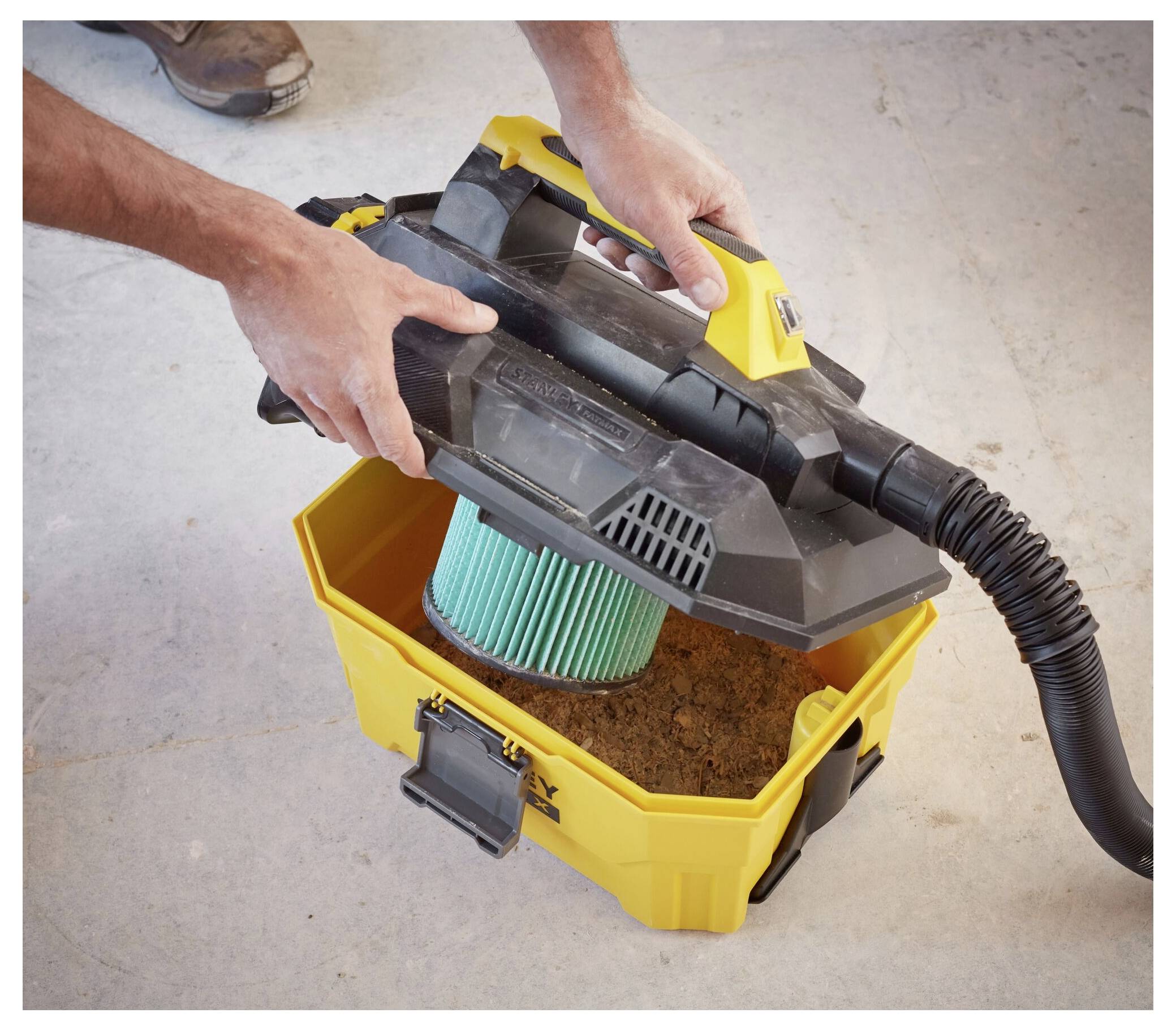 Hands holding a handheld vacuum placed above a yellow bucket filled with dirt, showcasing the vacuum's removable filter.