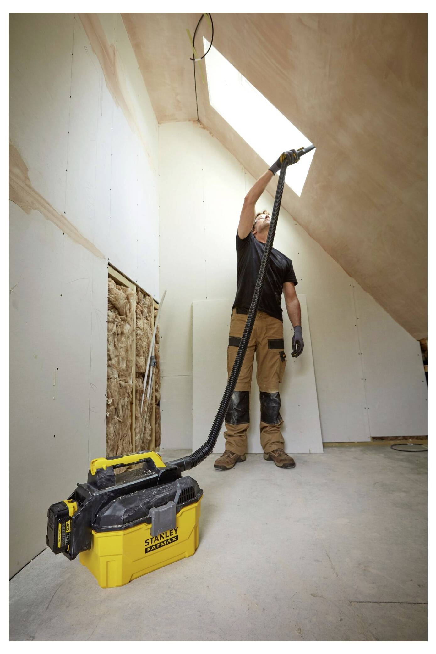 A person installing drywall in a room with sloped ceilings, using a shop vacuum cleaner to clear dust and debris, with natural light from a skylight.