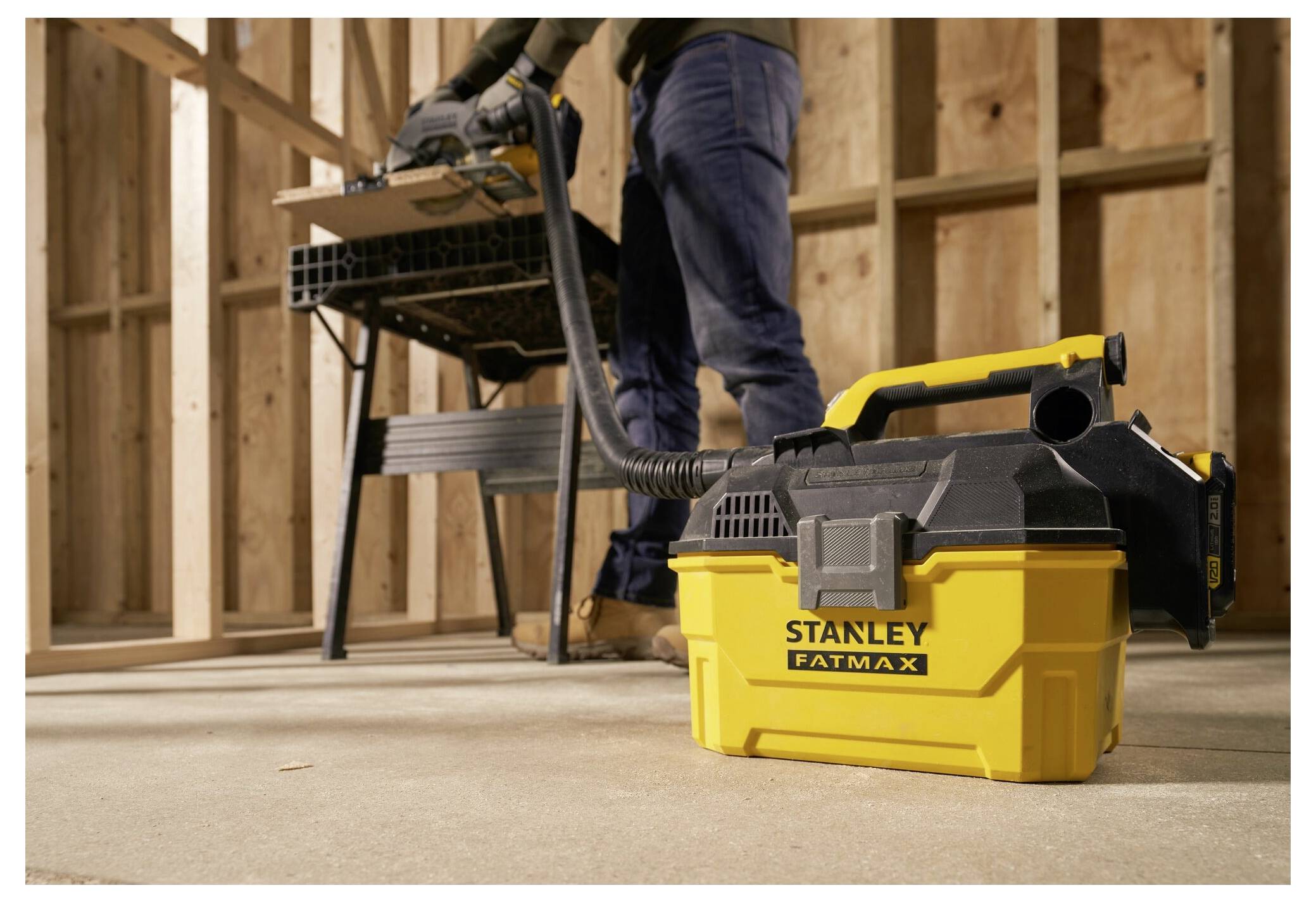 A vacuum cleaner labeled 'Stanley Fatmax' is on the floor with a hose connecting to a miter saw on a workbench. A person stands nearby.