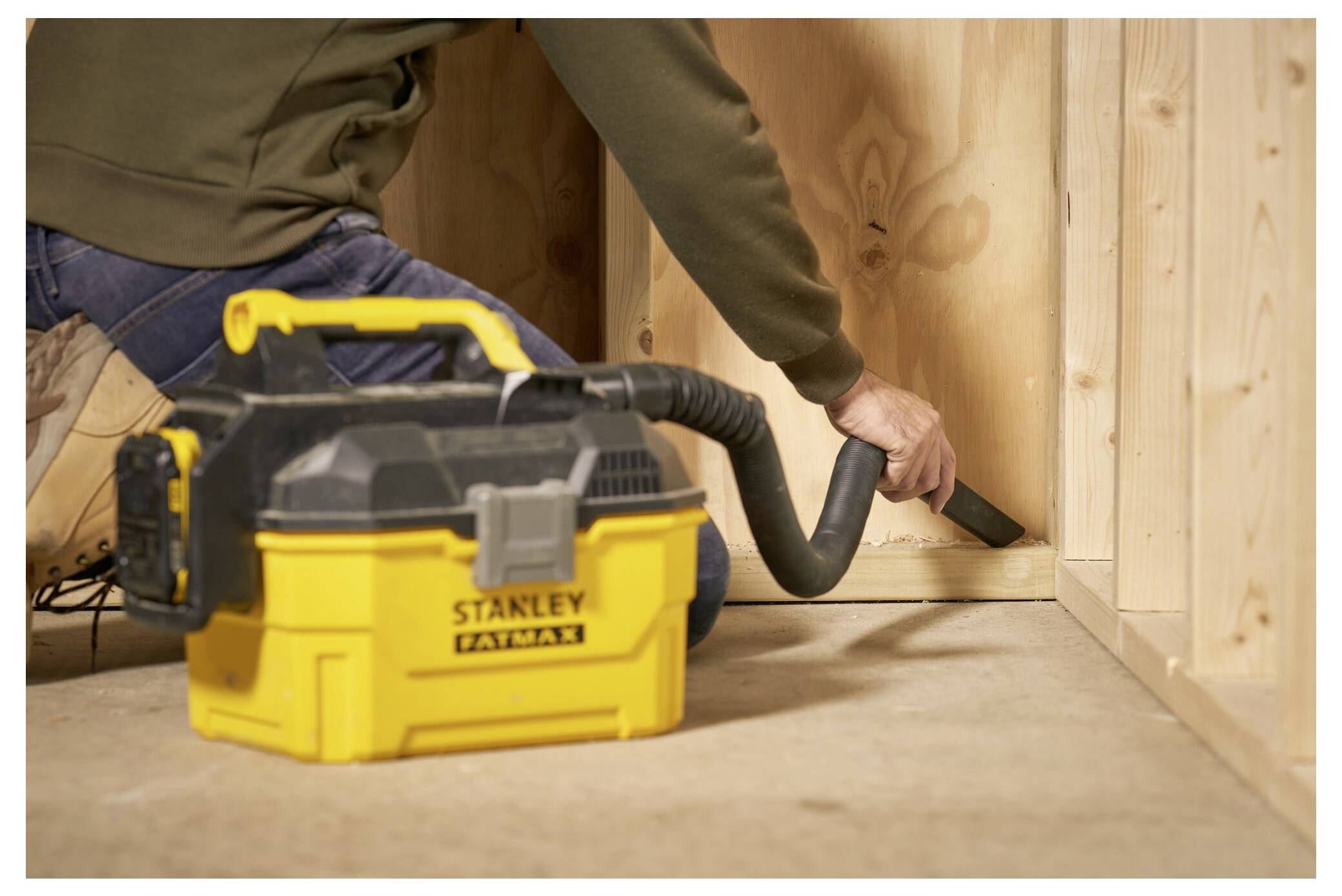 A person uses a portable yellow vacuum cleaner to clean dust from a corner of a wooden structure.