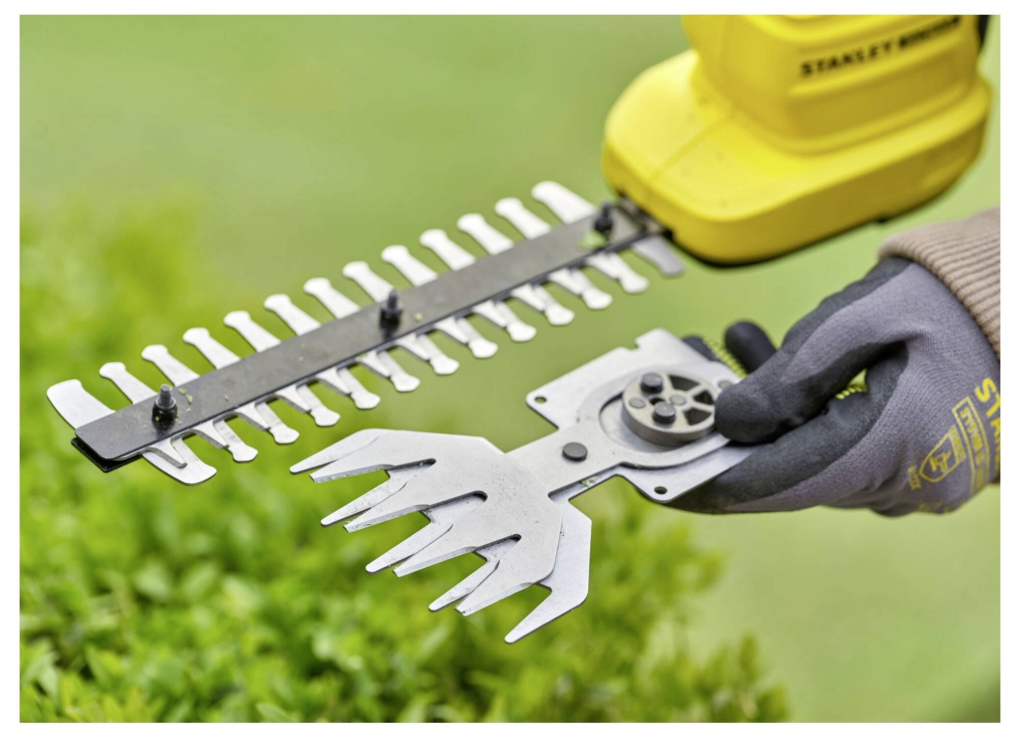 A person wearing gloves holds interchangeable blade attachments for a yellow hedge trimmer, with green bushes in the background.