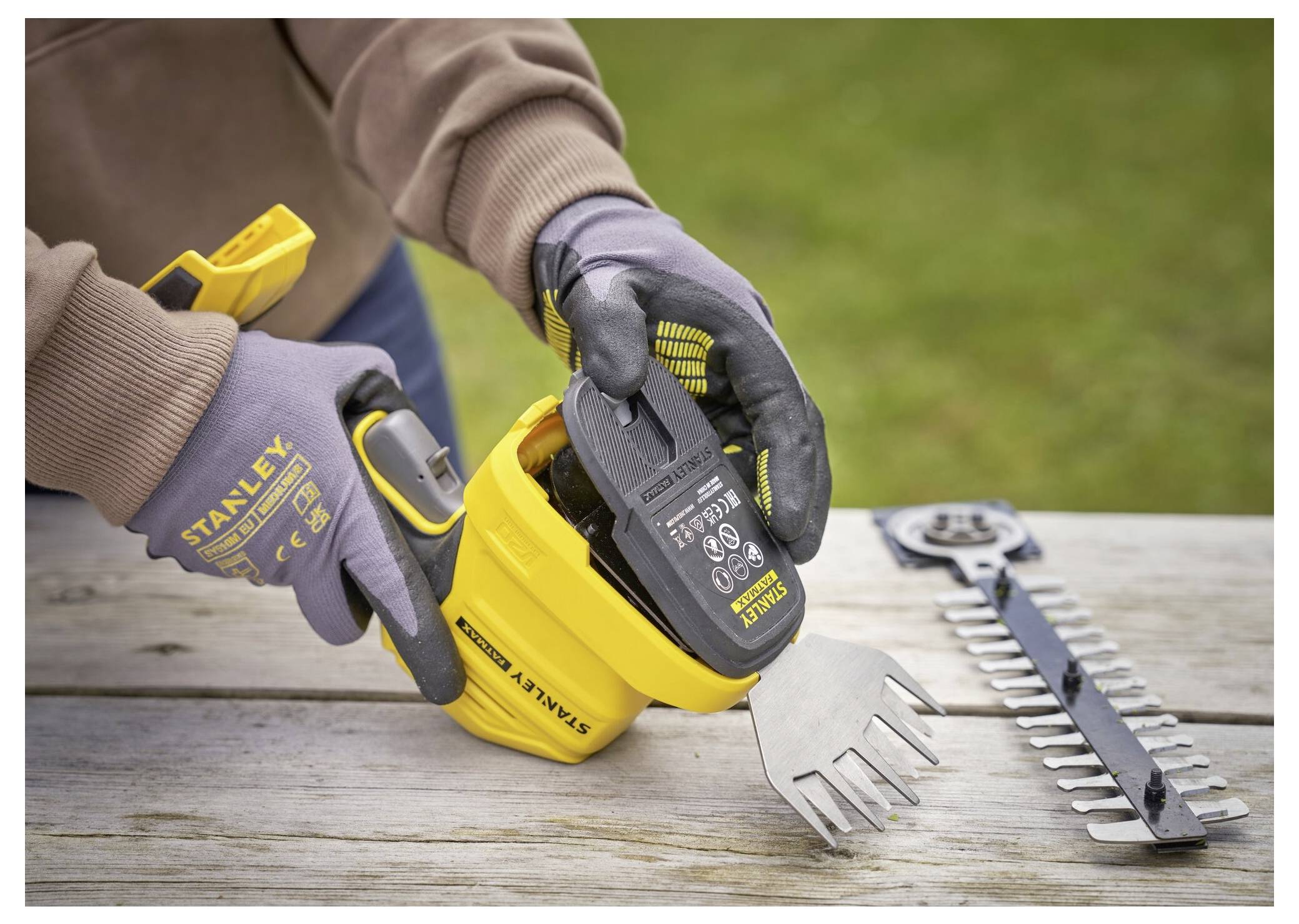 A person in gloves is attaching a battery to a yellow gardening tool on a wooden table, with a hedge trimmer attachment nearby.