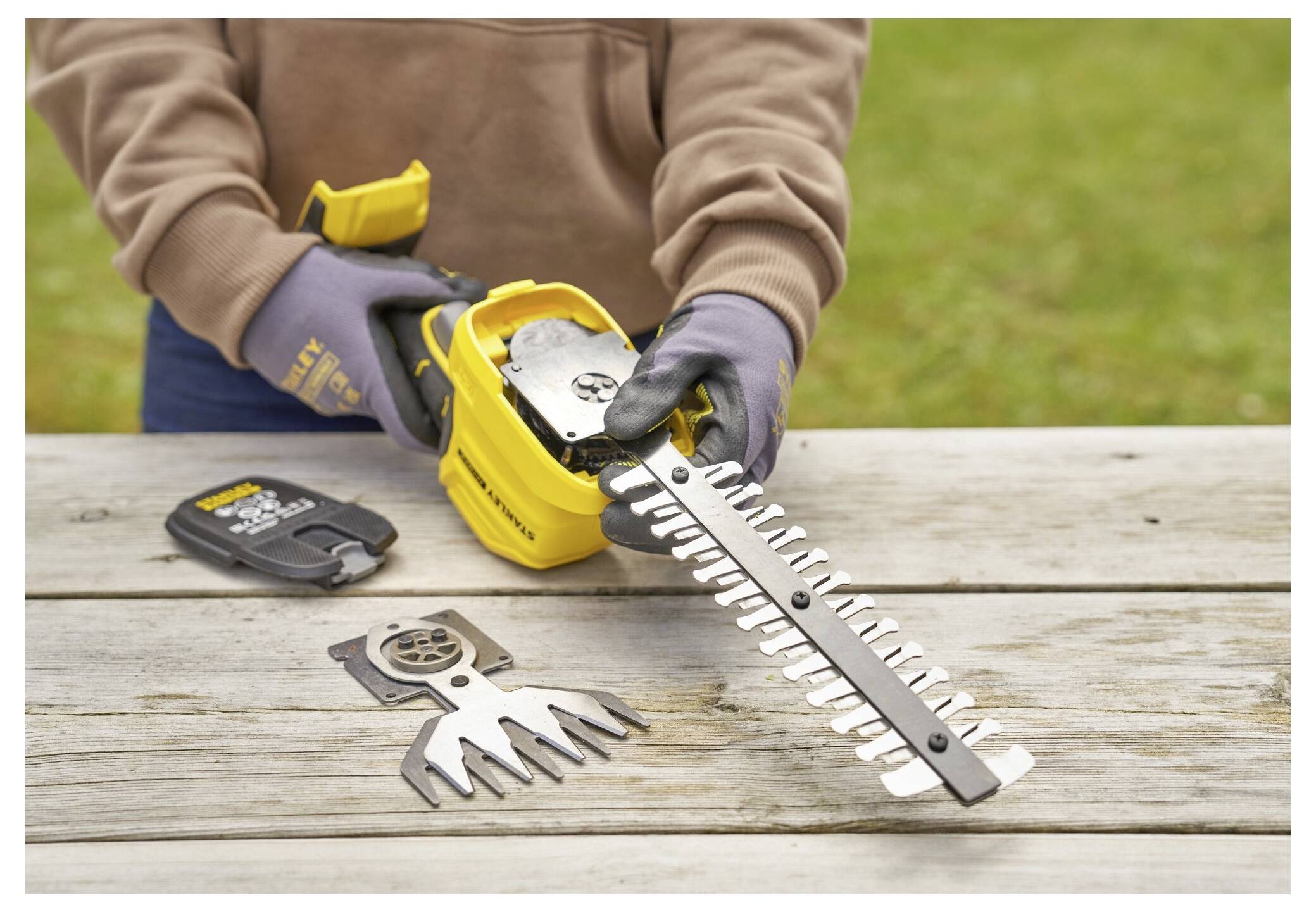 A person using gloves changes the blade of a yellow cordless brush cutter on a wooden table, with an additional blade beside it.
