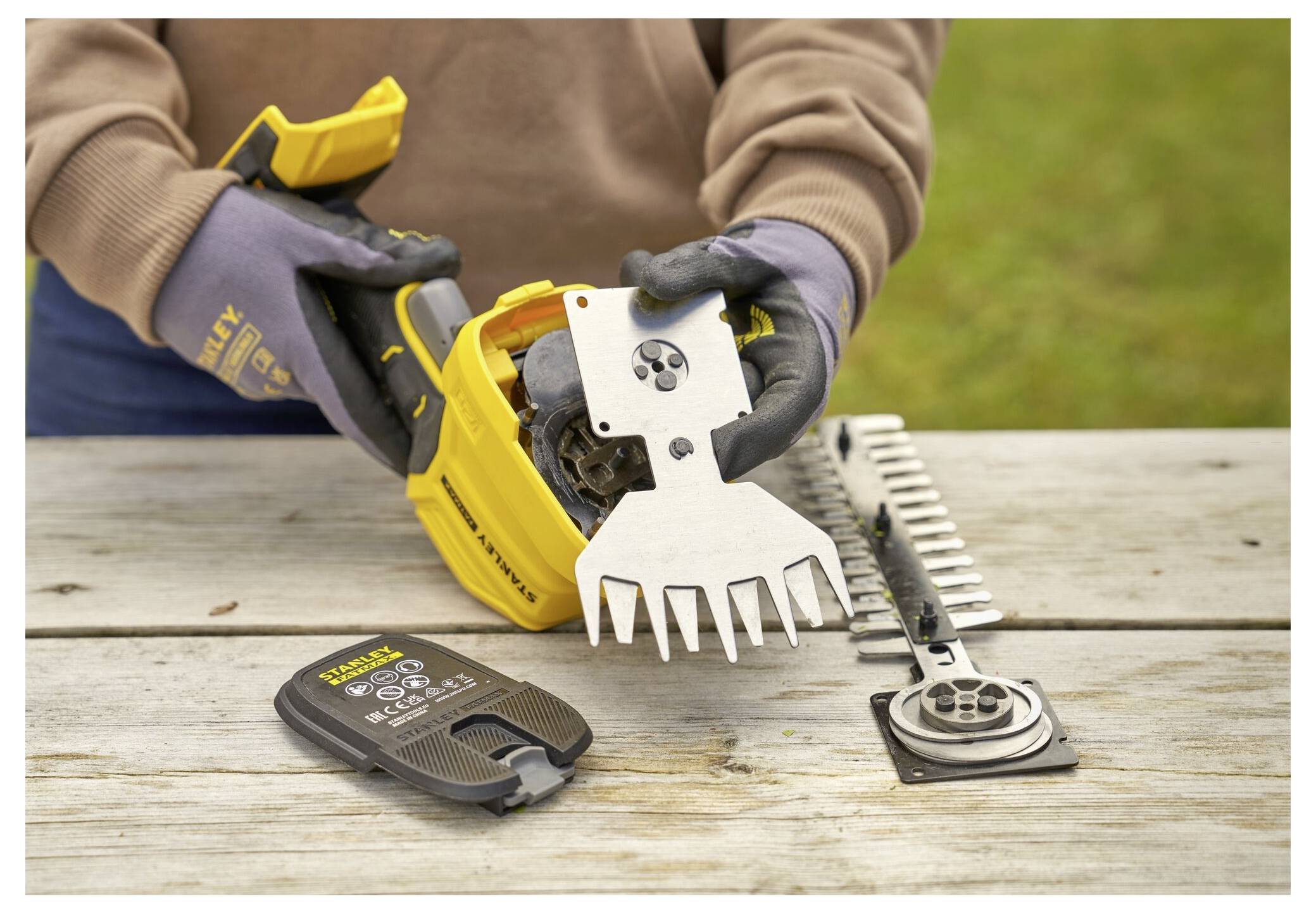 A person changes a blade on a yellow hedge trimmer, with additional blades and a charger visible on a wooden table outdoors.
