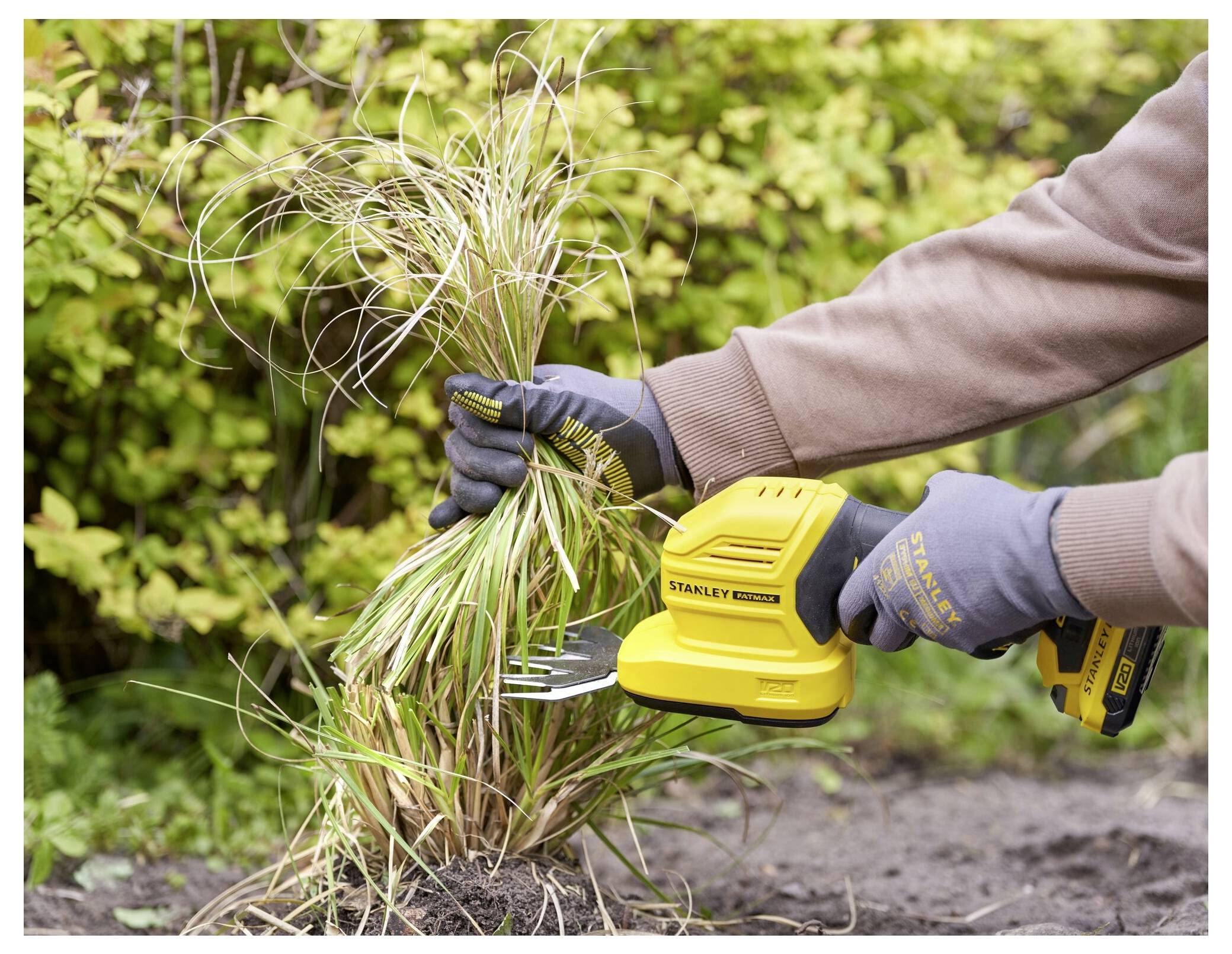 A person wearing gloves trims grass with a yellow electric tool in a garden, surrounded by green foliage.