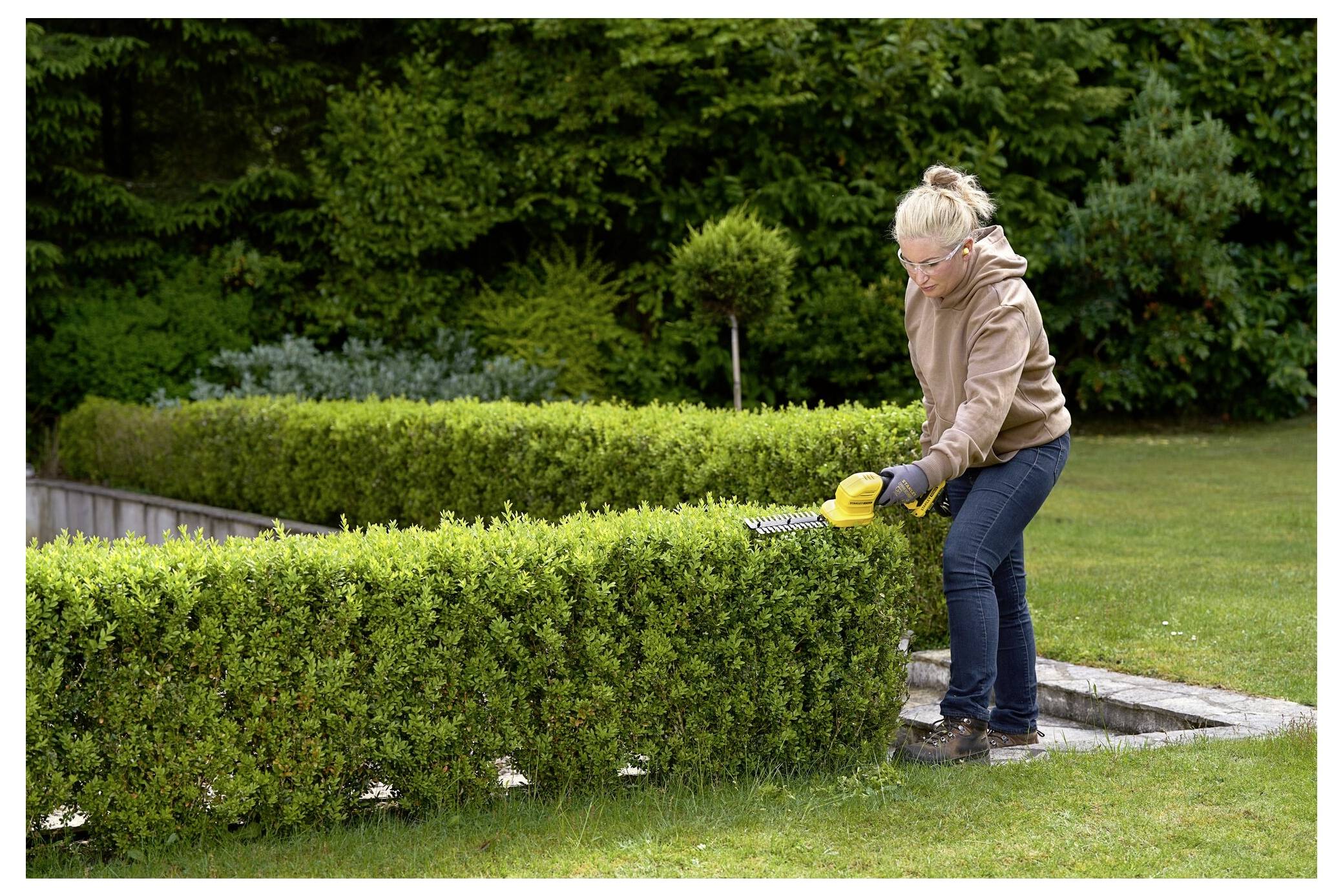 A person trimming a neatly shaped hedge in a garden, wearing gloves and a protective jacket, surrounded by lush greenery.