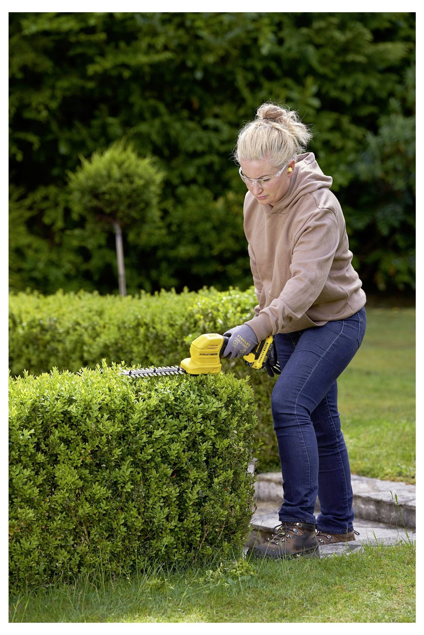 A person trimming a hedge with an electric hedge trimmer in a garden setting, wearing safety glasses, gloves, and casual clothing.