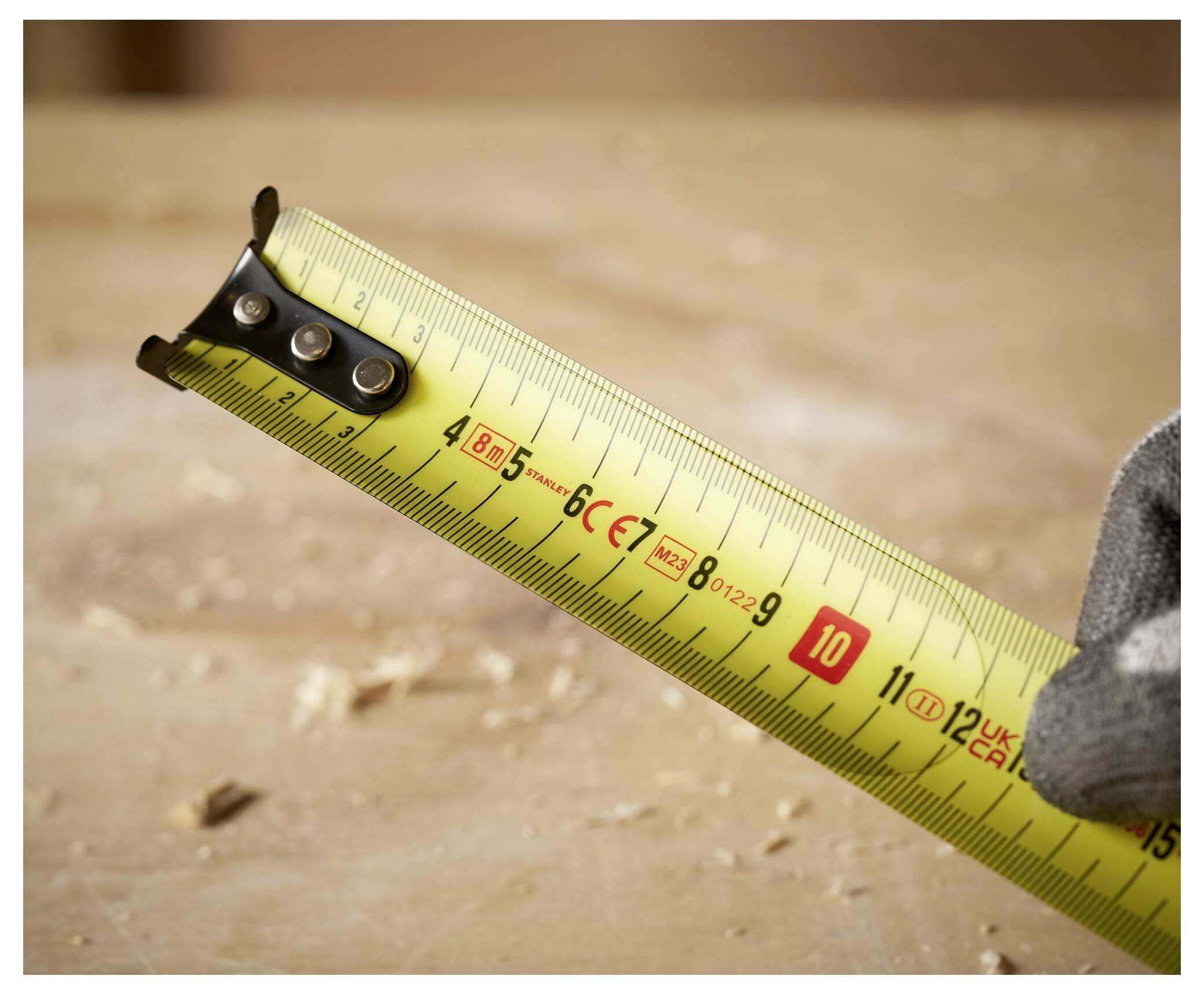 A close-up of a yellow tape measure extended across a wooden surface with sawdust, showing measurements in centimeters and millimeters.