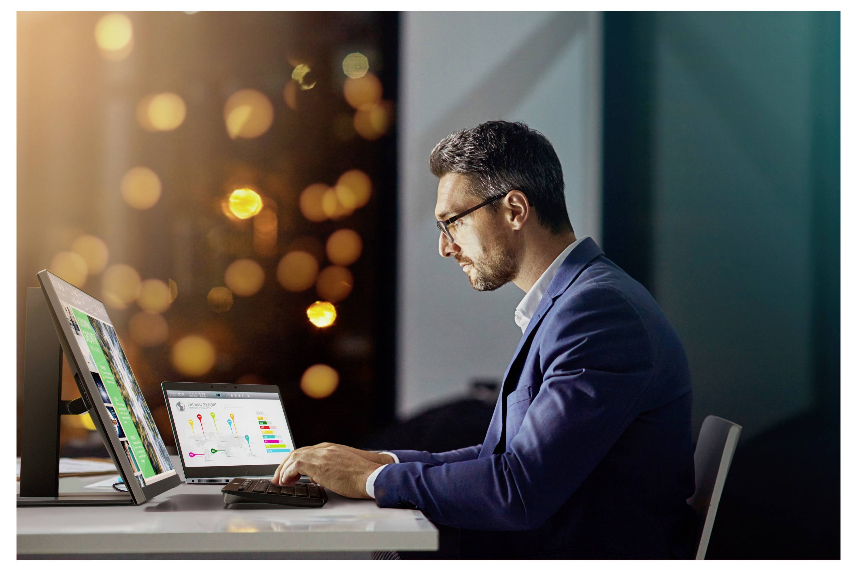 A man in a suit works on a computer displaying graphs and data. The background is blurred with city lights visible.