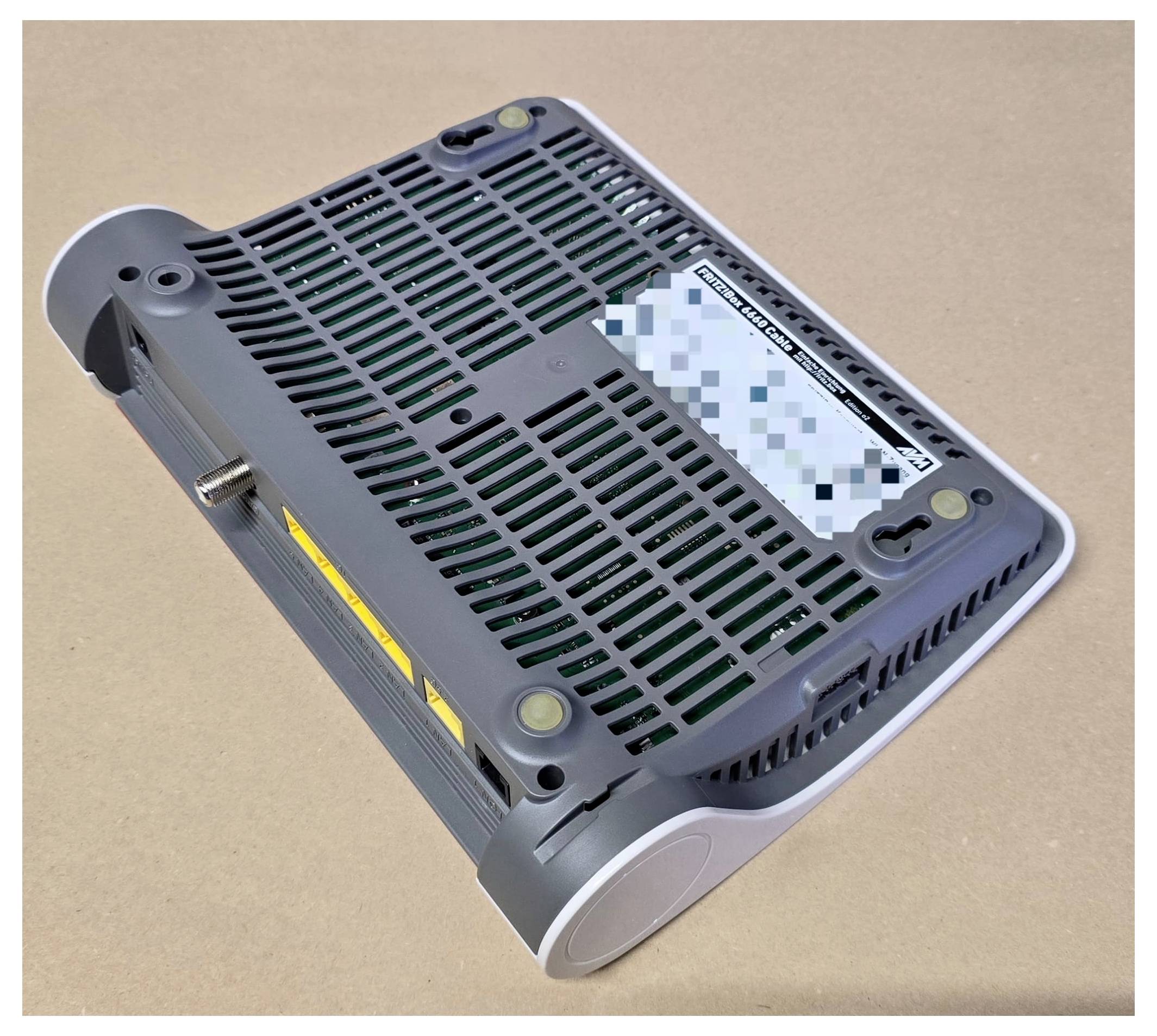 The image shows the underside of a black electronic device on a brown surface, featuring ventilation slots and a partially visible label.