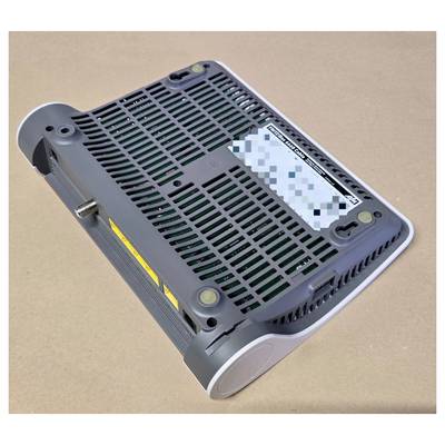 The image shows the underside of a black electronic device on a brown surface, featuring ventilation slots and a partially visible label.
