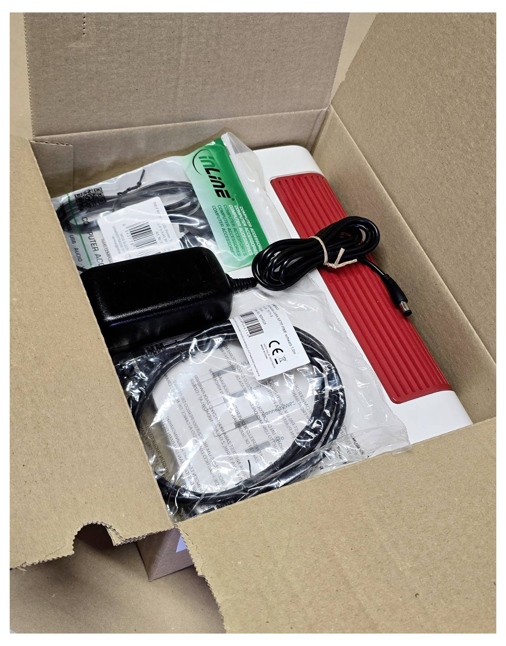 An opened cardboard box contains an electronic device with a red handle, wrapped cables, a power adapter, and an instruction manual.