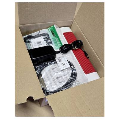 An opened cardboard box contains an electronic device with a red handle, wrapped cables, a power adapter, and an instruction manual.