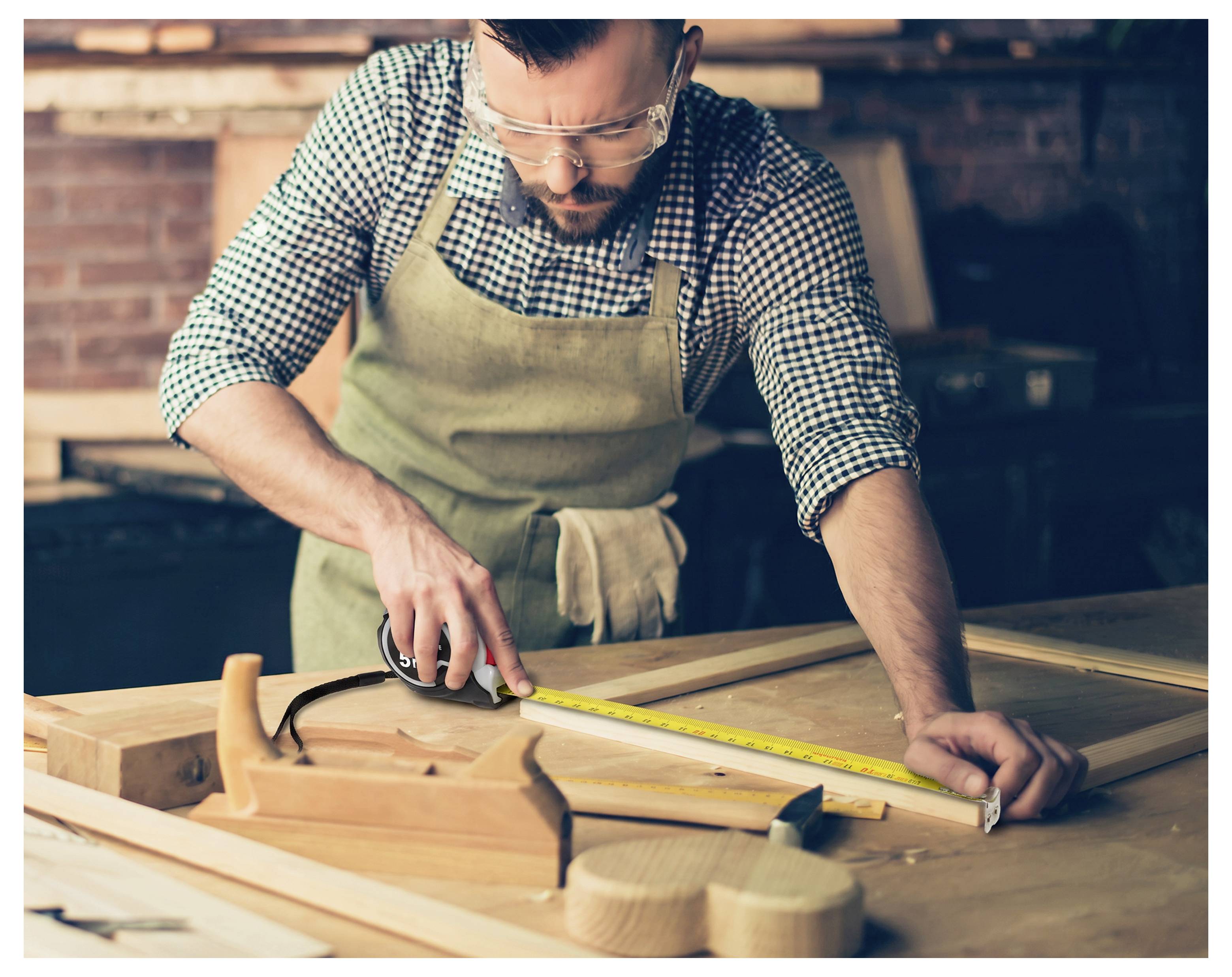 Woodworker in an apron measures a wooden board with a tape measure at a workbench in a workshop, with hand tools nearby.