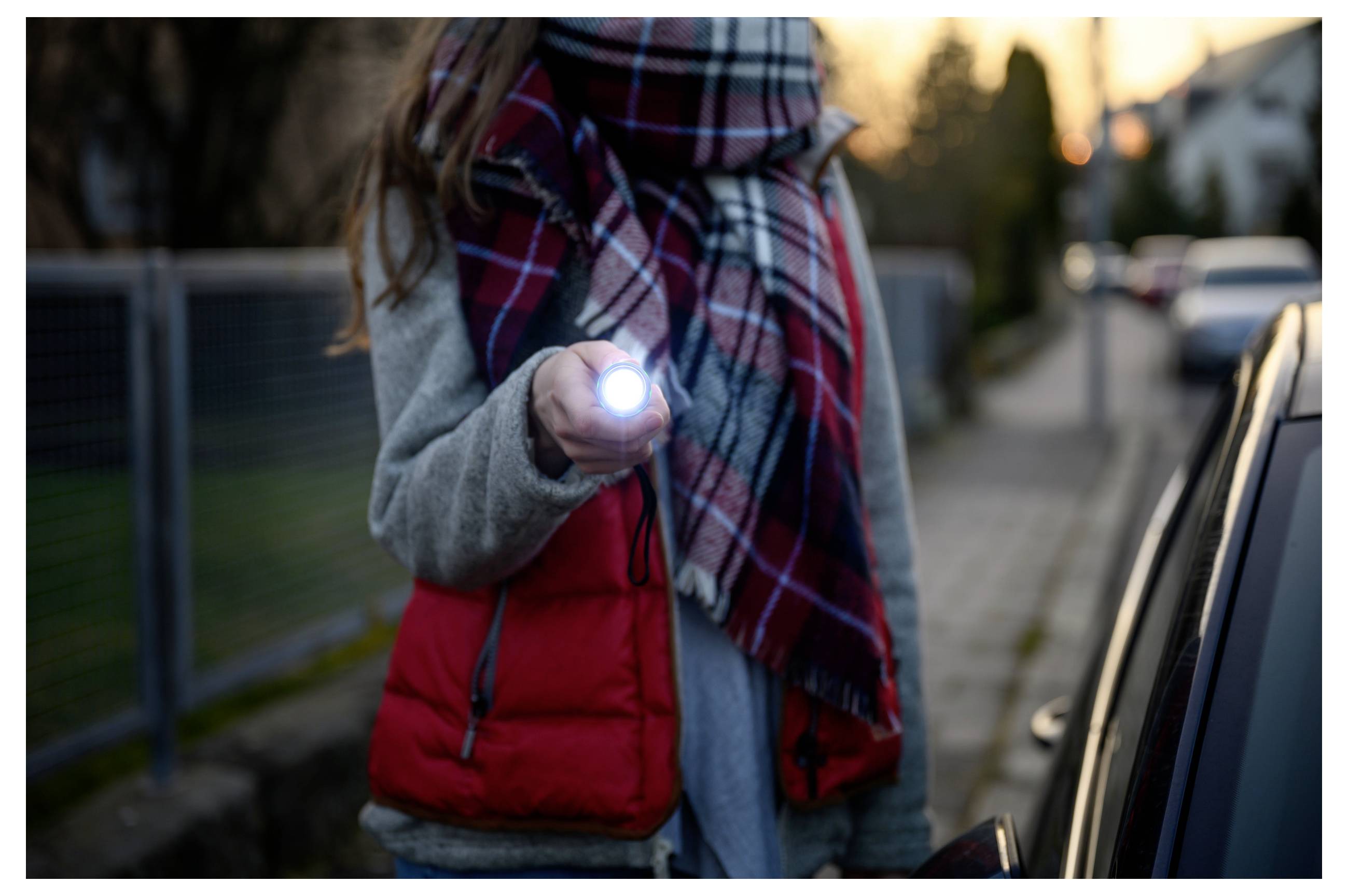 Person on a sidewalk shining a flashlight toward a car at dusk, suggesting checking or signaling near a parked vehicle.