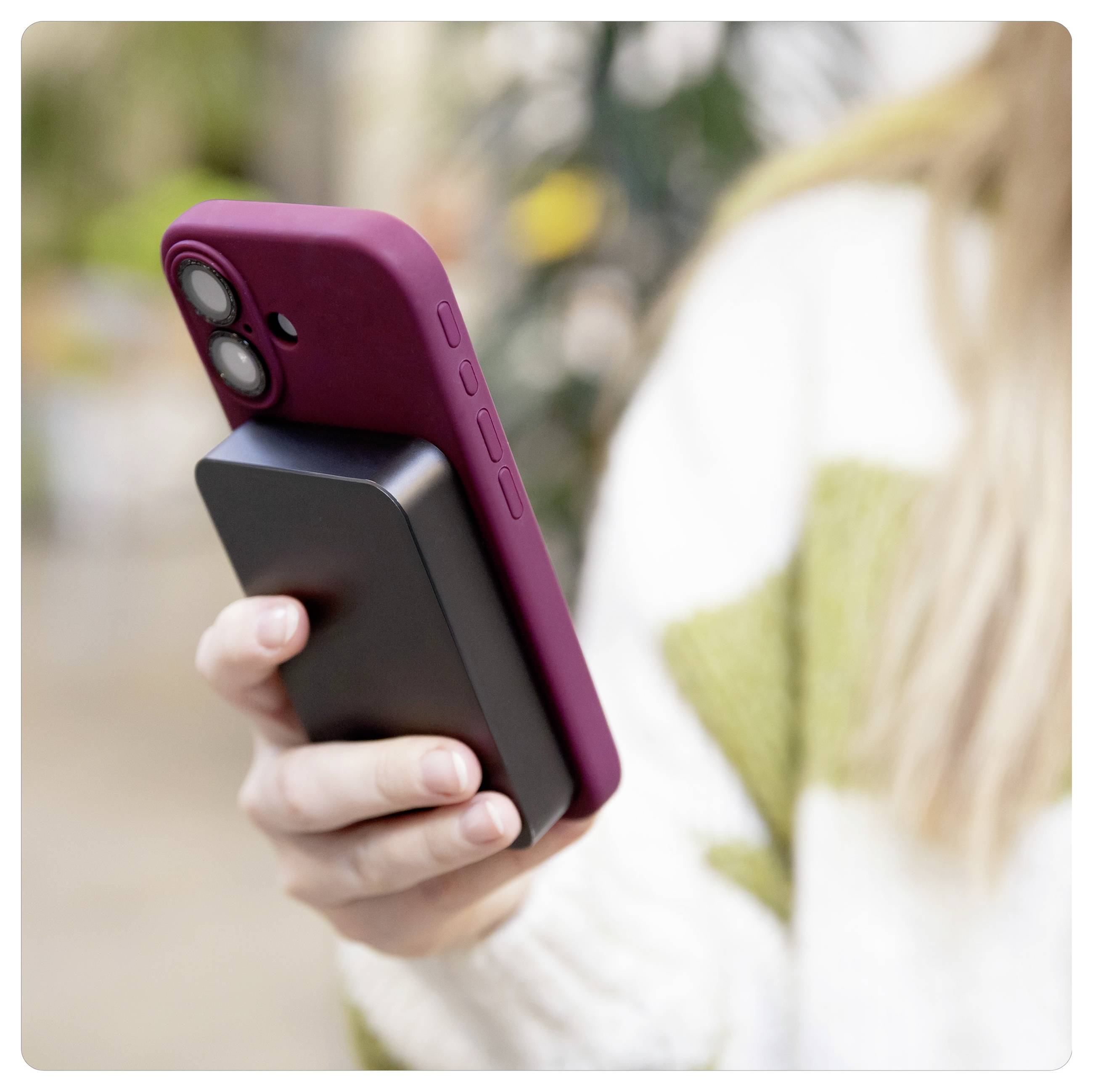 Hand holding a smartphone with a magenta case and attached black battery pack outdoors.